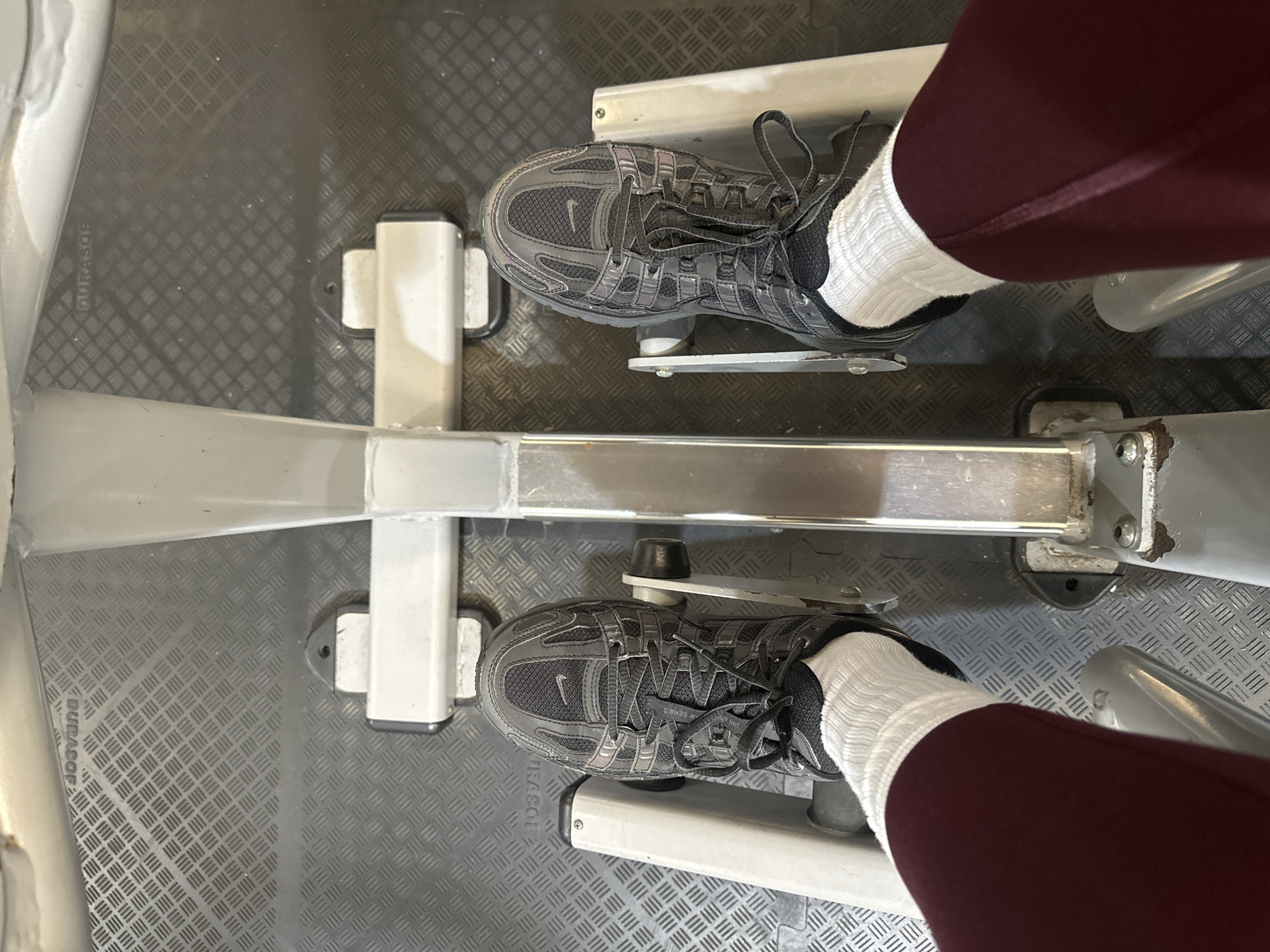 A first-person, downward perspective captures a person's lower legs and feet on a grey metal exercise machine within a fitness facility in Tunbridge Wells, United Kingdom. The individual is wearing dark red athletic leggings, white ribbed ankle socks, and dark grey Nike athletic shoes with black laces. Their feet are placed on the adjustable, textured footplates of the machine, which features a central structural beam. The exercise machine, consistent with a rowing machine or a leg press, rests on dark grey anti-slip gym flooring. This matting displays a raised diamond pattern, with the inverted brand name "DURASOF" visible in the upper background.