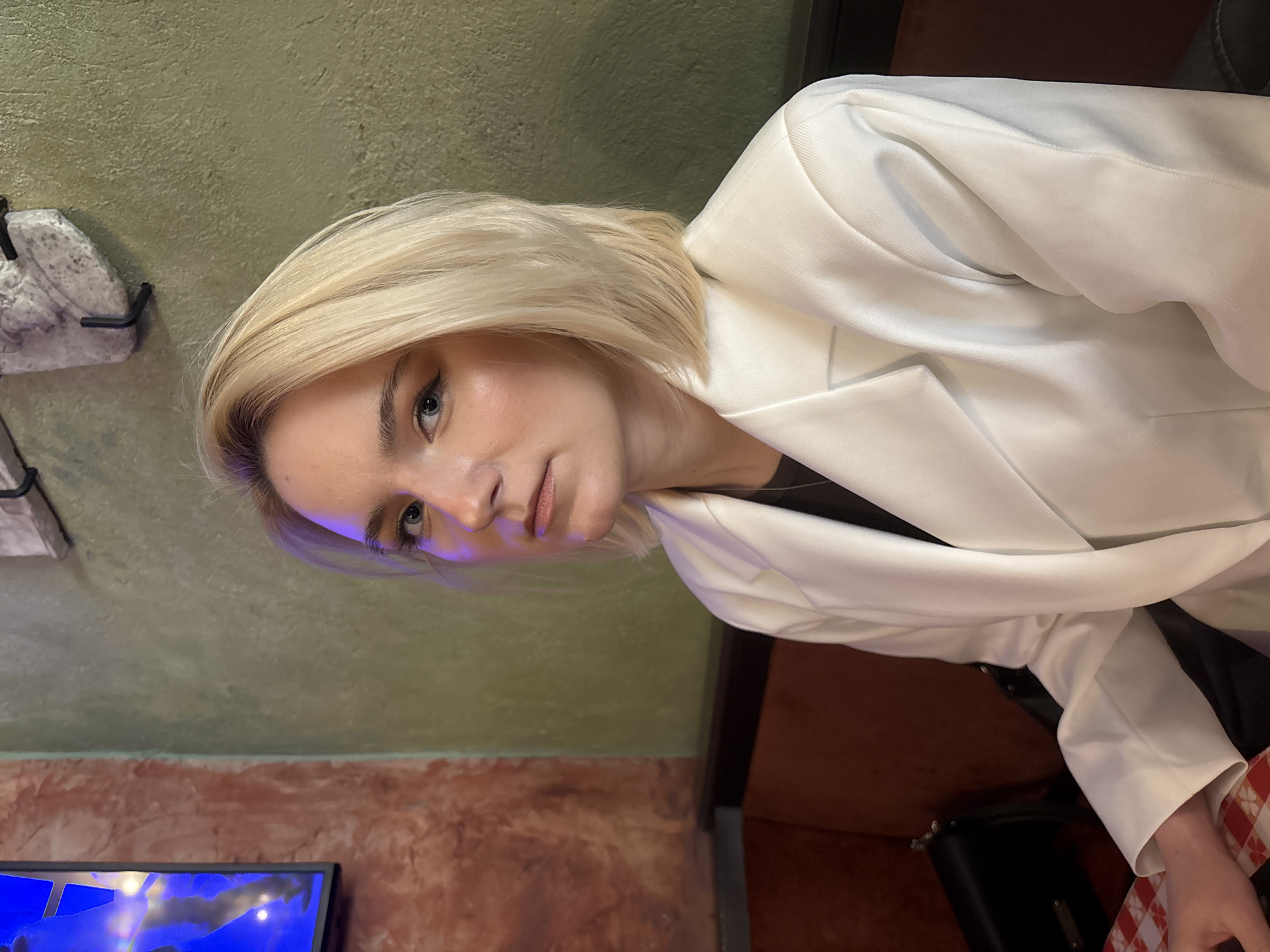 The image captures a headshot and upper body view of a young woman with short, straight blonde hair, framed against an indoor background. She has light-colored eyes, defined eyebrows, and appears to be wearing subtle eyeliner and lip makeup. Her expression is calm and direct, as she looks towards the viewer. She is dressed in a crisp white blazer worn over a dark top.
The setting appears to be an interior space, likely an establishment such as a cafe, bar, or restaurant in Saint Petersburg, Russia. The background features two distinct sections of textured walls: an upper portion of muted, olive-green and a lower section of reddish-brown/terracotta. A horizontal line separates these two wall textures.
On the left side of the frame, a decorative, rough-textured grey object, possibly made of stone or concrete, is partially visible, featuring dark metal brackets or hooks. In the bottom left corner, part of a screen, possibly a television or monitor, displays vibrant blue and white abstract colors. Further to the right, at the bottom, a black bag or object is visible, next to a piece of fabric with a red and white checkered pattern, which could be part of a tablecloth or another item.
The lighting suggests an indoor, artificial setting, possibly in the evening or at night. A subtle purple or blue hue is noticeable on the woman's face and hair, which might be a reflection from the screen or a specific lighting choice in the venue. There is no visible text in the image. The woman is the central subject, posing for the photograph.