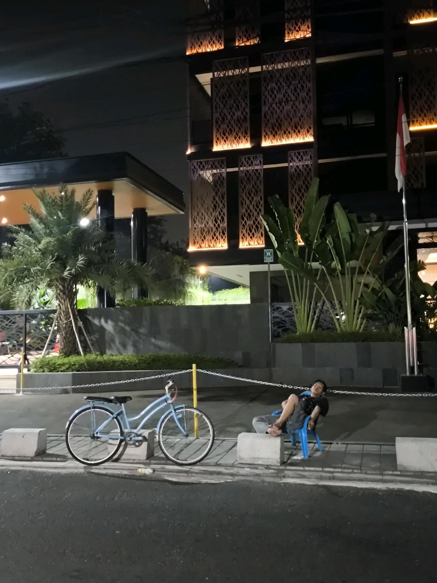 A man is relaxing on a blue plastic chair in front of a building at night. He is wearing a black t-shirt and gray pants, with his legs crossed and his head tilted back. A light blue bicycle is parked next to him, leaning against concrete blocks. The building has decorative metal screens illuminated from behind with warm orange light. A flag pole with an Indonesian flag is visible to the right of the building. The scene is set on a paved area with some greenery and trees, suggesting an urban environment. This is likely in the city of West Jakarta, Indonesia, given the Indonesian flag.