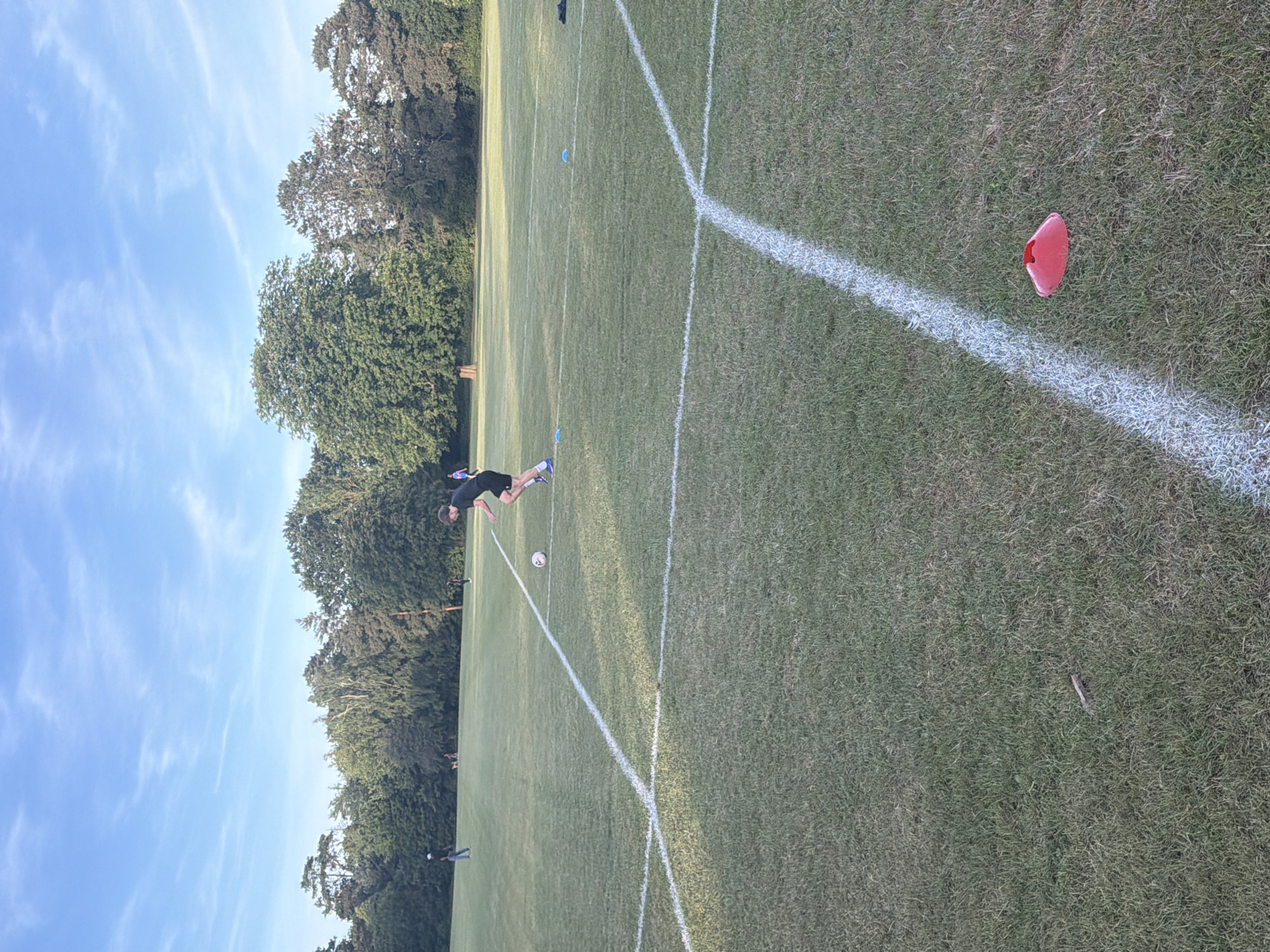 The image depicts an outdoor grassy field, identified as being in Maidstone, United Kingdom, under a bright blue sky with sparse, wispy white clouds. The central action involves a young male, positioned on the mid-right, wearing a dark t-shirt, dark shorts, and blue athletic footwear, actively dribbling a white and black football with his right foot. He is moving from right to left across the frame.
The field is marked with several white lines, two of which prominently intersect in the mid-foreground, forming a corner of a marked area. A red flat training cone is visible in the bottom-right corner, adjacent to one of these white lines. Further back on the field, two blue flat training cones are discernible.
In the mid-ground and background, a dense line of mature green trees forms the horizon. Several other individuals are present on the field: one person in a dark t-shirt and light trousers is standing towards the far left, near the tree line, while other smaller figures are visible further in the background near the trees. The grass appears well-maintained but shows some natural variations and slight wear. The lighting suggests a clear day, possibly late afternoon or early evening, casting soft shadows.