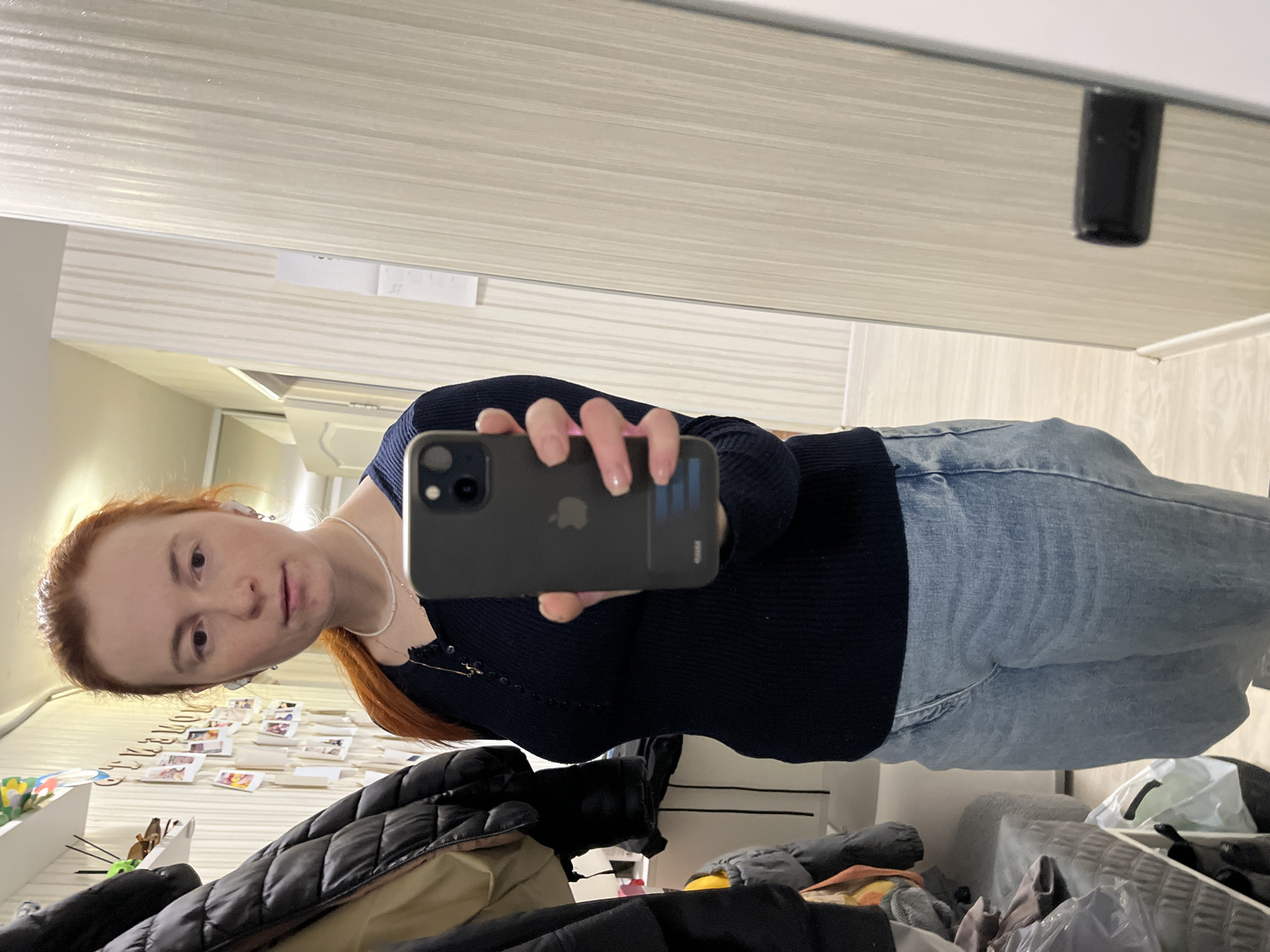 A woman with red hair is taking a selfie in what appears to be a room reflected in a mirror. She's wearing a dark, possibly navy, sweater and light blue jeans. She holds a black phone in front of her face, partially obscuring it. The room contains furniture, scattered items, and photos on the wall. Based on the location Chaikovsky, Russia, the environment would be a typical residential interior for that area.