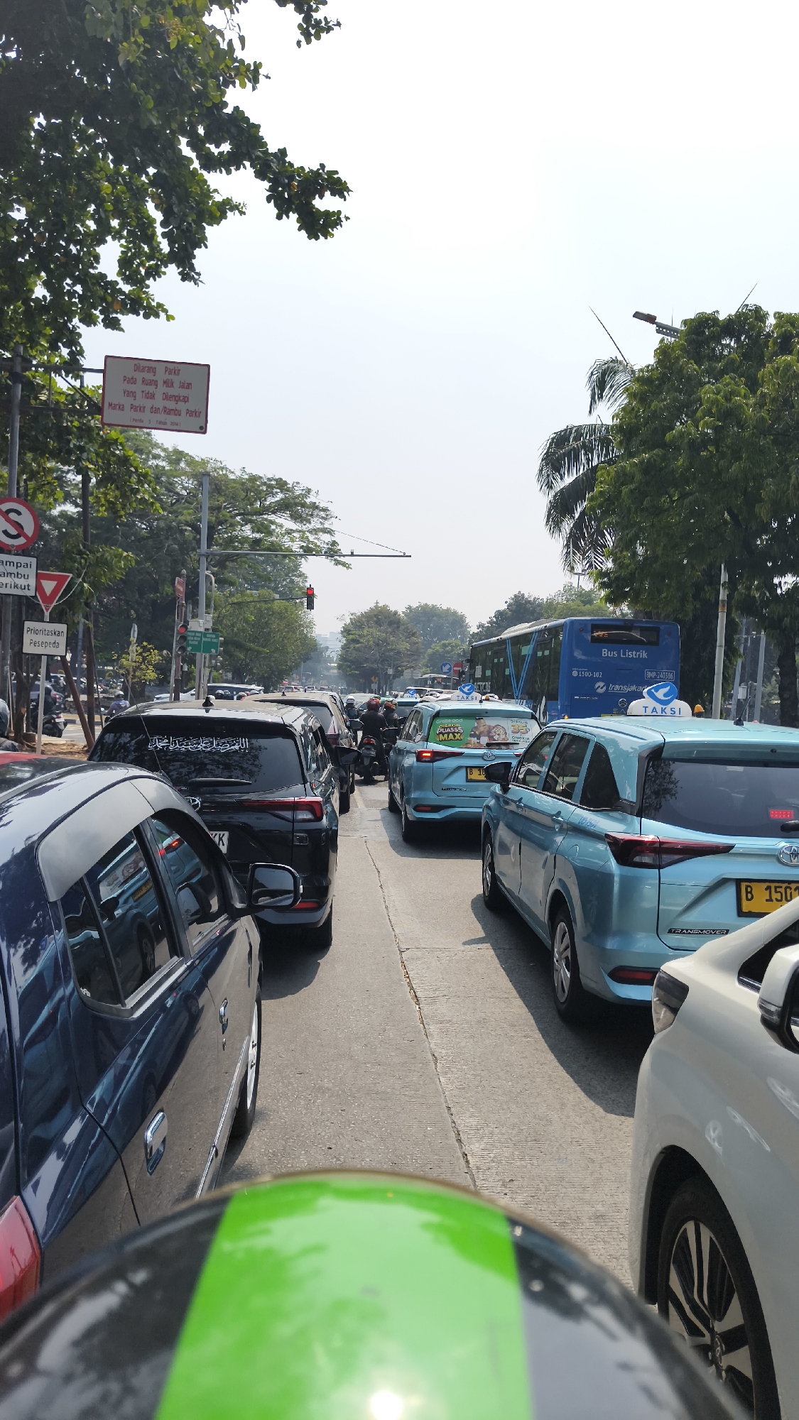 The image shows a congested city street in South Jakarta, Indonesia. The perspective is from a motorcycle, looking forward at a line of cars, including a dark SUV in the foreground, several light blue taxis, and a blue electric bus in the background. Traffic lights are visible and appear to be red, indicating a standstill. Road signs are present, including one that states "DILARANG PARKIR" (No Parking) and another indicating "Prioritaskan Pesepeda" (Prioritize Cyclists). Trees line the street, and the sky is overcast. The overall scene depicts heavy urban traffic.