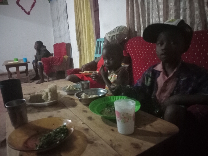 A family sits around a wooden table laden with food. On the table are plates of what appears to be mashed root vegetables, possibly yams or cassava, alongside green leafy vegetables, and a drink in a plastic cup. A young boy in a hat sits near the camera with a younger child beside him. Behind them sits a woman. In the background, a man is sitting on a chair. The scene appears to be a family meal in a modest home.