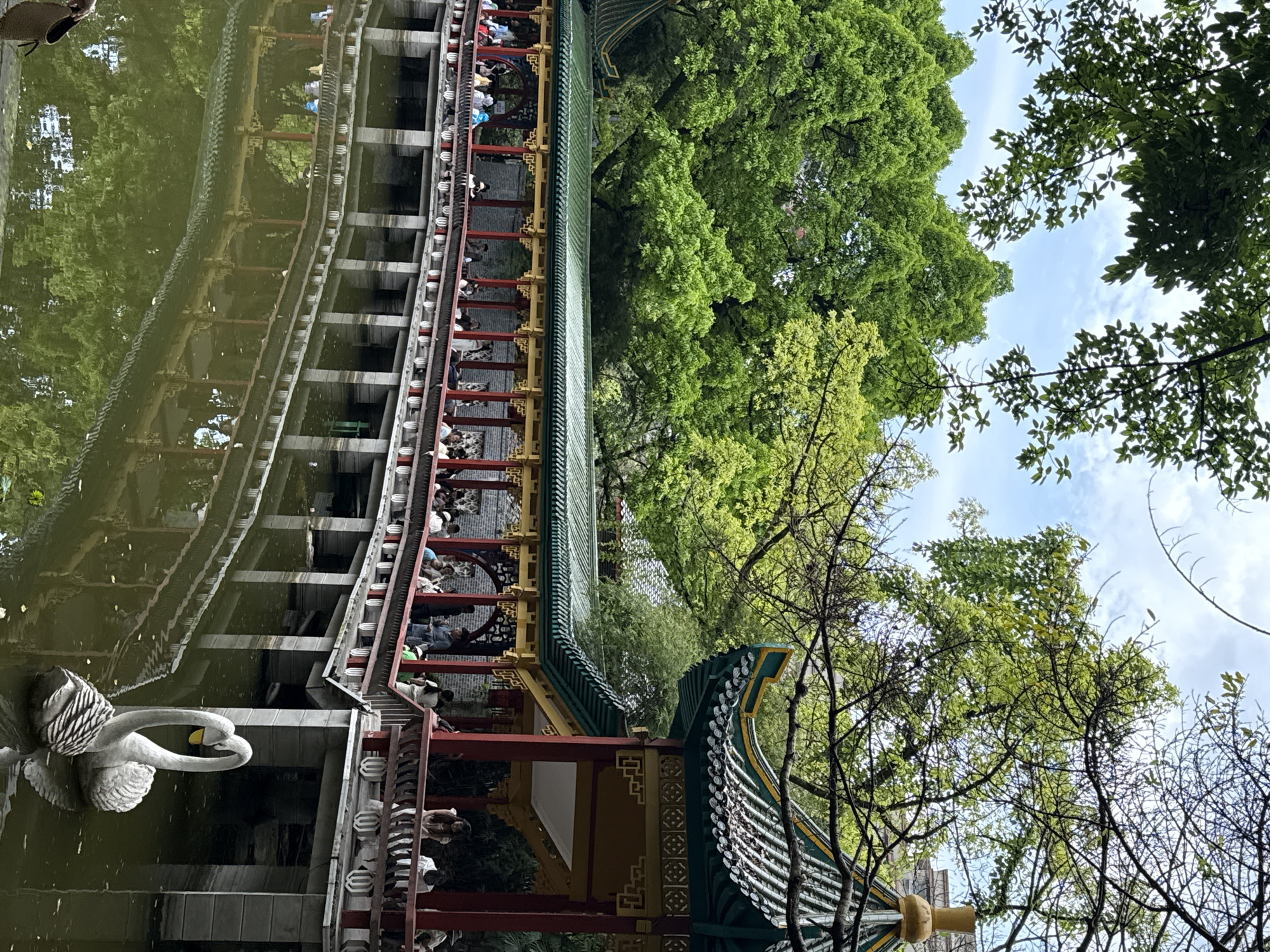 The image shows a traditional Chinese garden scene, possibly in the Yuzhong District of China. A long, covered walkway with red pillars and a green tiled roof runs alongside a body of water. People are seen walking or standing on the walkway. The water reflects the walkway and surrounding trees. A white swan statue floats on the water near the walkway. Trees with dense green foliage are visible in the background, partially obscuring the sky. The architecture appears traditional, with ornate details.