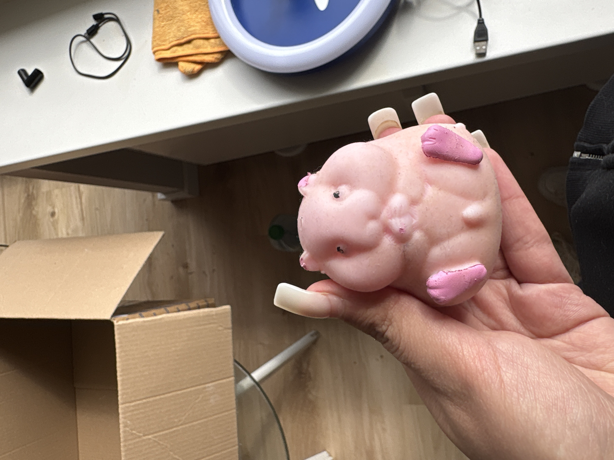A close-up shot from an elevated angle depicts a person holding a pink, toy-like pig with painted hooves. The individual's hand exhibits long, square-shaped nails. In the background, a partially open cardboard box is visible. The setting includes a surface with various items: a small black heart-shaped object, a black wired accessory, an orange cloth, a blue circular object with a white center, and a USB cable. The locale appears to be an indoor space, and without additional details, it is impossible to confirm if the image was captured in Mönchengladbach, Germany.