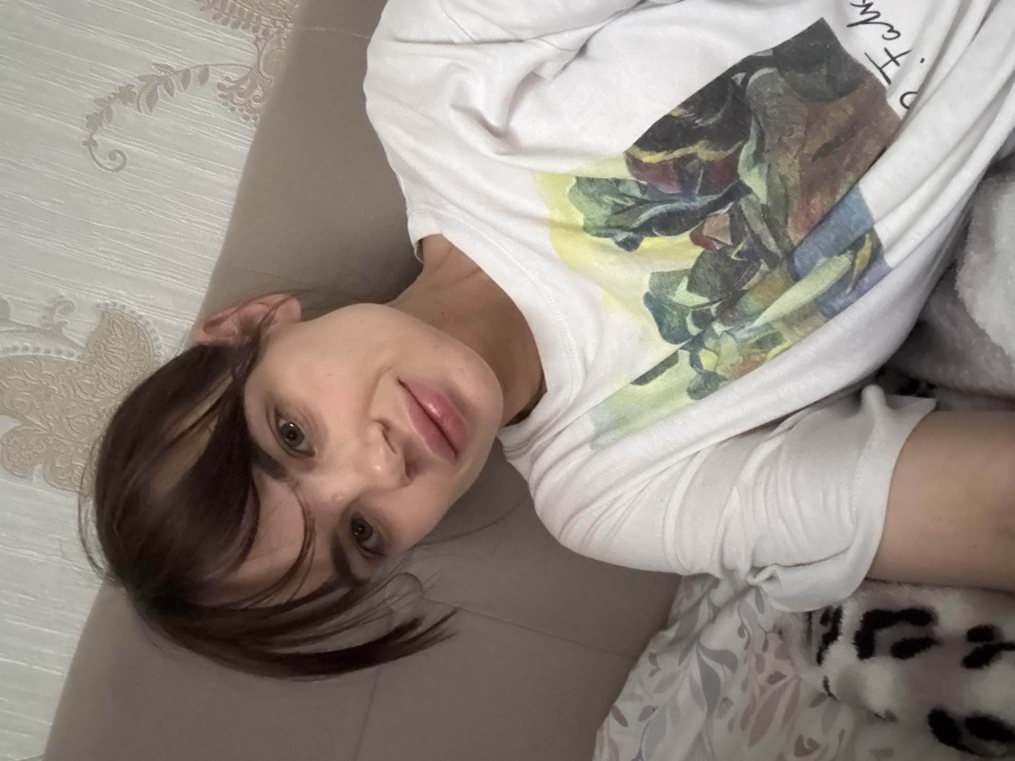 A woman lies on a bed, wearing a white t-shirt with a painting. She has brown hair with bangs and fair skin. The bedding is patterned, with a mix of floral and animal prints. The background includes a textured wall. The location cannot be determined from the image.
