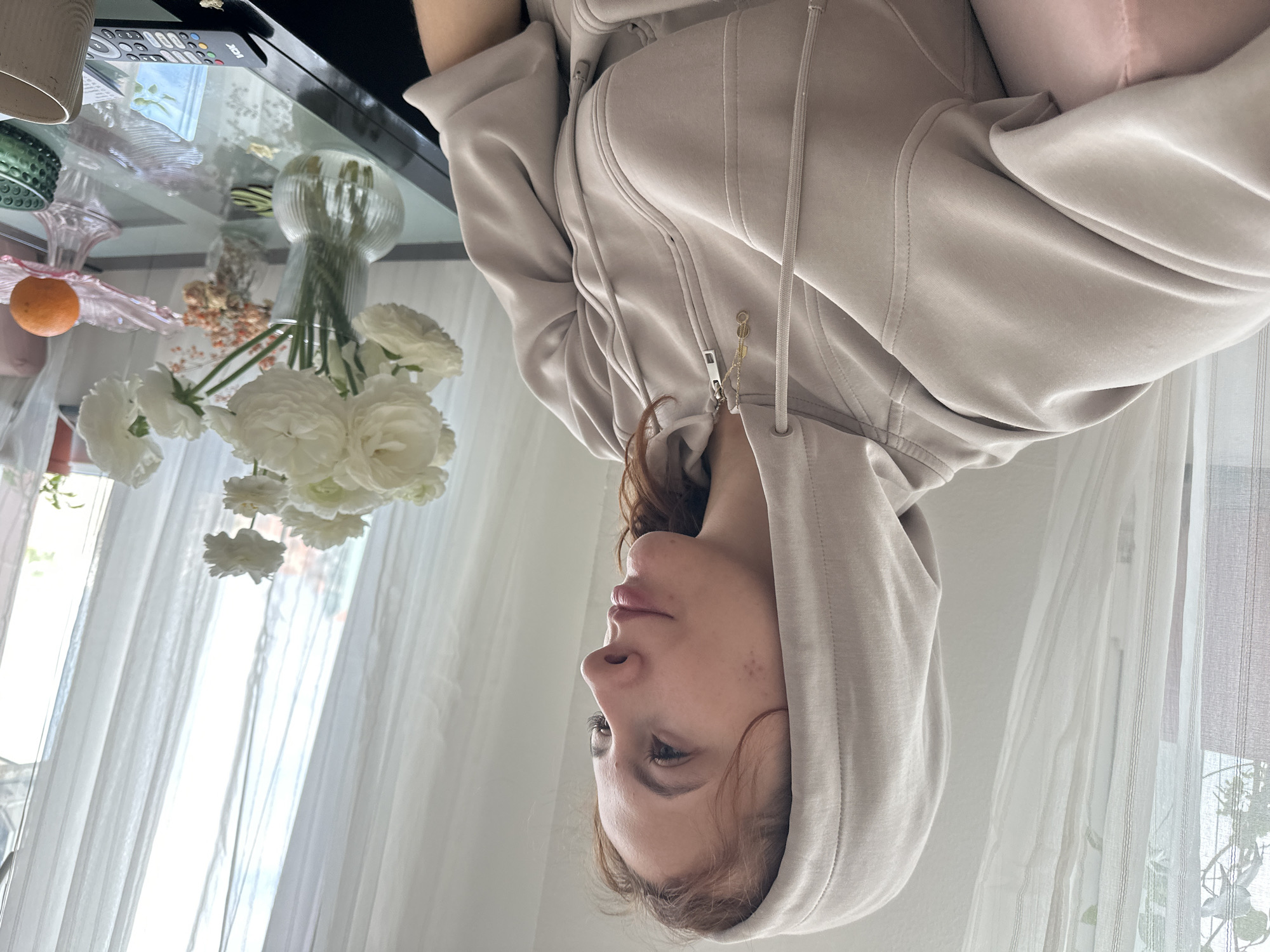 This portrait-style photograph captures a young person, likely female, in an indoor setting in Novi Sad, Serbia. The person is seen in profile, wearing a light beige or tan hooded sweatshirt with the hood pulled up. Her reddish-brown hair is visible beneath the hood, and her face is turned slightly upwards and to the left, displaying a calm and contemplative expression. A small golden charm, possibly spelling "LOVE," is attached to the zipper pull of her sweatshirt.
In the foreground and midground, a glass-top table is adorned with several decorative items. A prominent bouquet of white ranunculus flowers with green stems is arranged in a ribbed glass vase. A small orange fruit, resembling a tangerine or clementine, is placed on the table next to the flowers. Other objects include a green textured glass bowl or vase, a pink ruffled glass dish, and a light-colored ceramic cup or planter. A black remote control with various colored buttons, featuring the text "RCA," rests on the far left edge of the table.
The background reveals sheer white curtains, indicating a window that allows bright, diffused natural light to illuminate the scene, suggesting it is daytime. The overall atmosphere is serene and domestic, likely a living room or dining area. The soft lighting enhances the calm mood conveyed by the person's expression.
