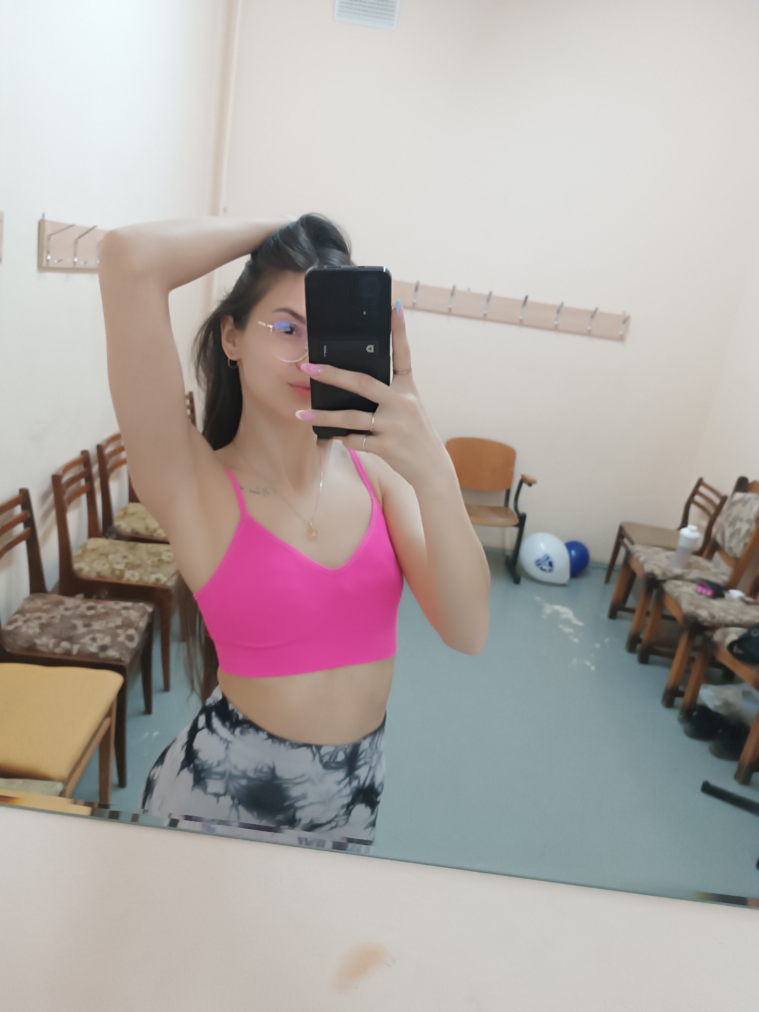 The image depicts a woman taking a selfie in what appears to be a changing room or locker room setting. The location is Unknown, Unknown.
**People:**
* One woman is visible, taking the selfie. She is wearing glasses, a pink sports bra, and gray and black tie-dye leggings. Her hair is long and dark.
**Objects:**
* A smartphone is held in the woman's hand, partially obscuring her face.
* The room contains rows of wooden chairs.
* There are clothes hooks mounted on the wall behind her.
* Two helmets and water bottles on the floor.
**Environment:**
* The room has a clean, institutional look. The walls are a light color, and the floor appears to be linoleum or tile.
* The scene is lit by artificial lighting.
**Action:**
* The woman is taking a selfie, holding her phone and posing with one arm raised.