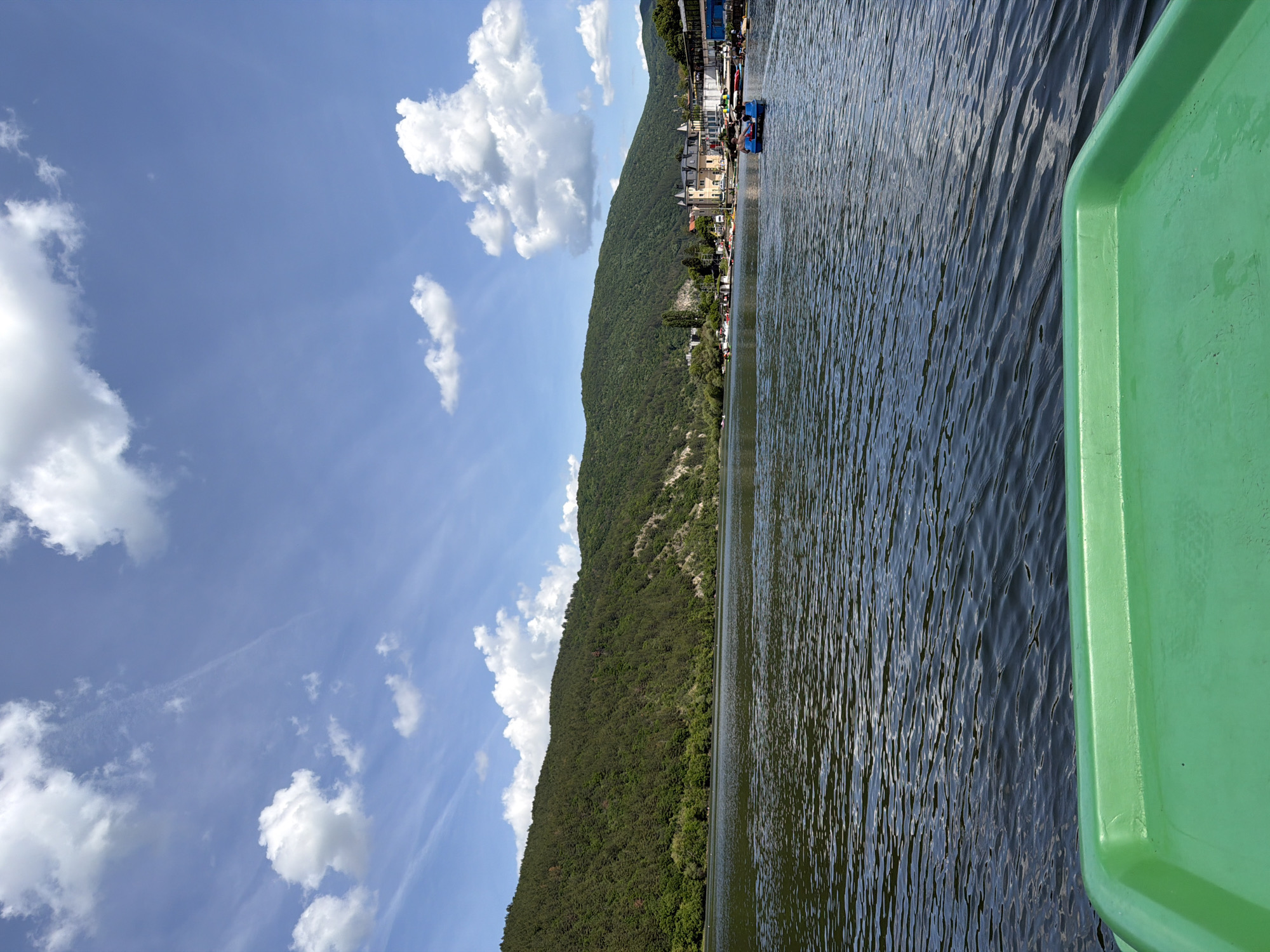 The photograph captures a scene on Lake Pancharevo, near Pancharevo, Bulgaria. The view is likely from a small boat on the lake, with a section of its green plastic edge visible in the lower right of the frame. The lake’s surface is rippled with visible water movement, reflecting the sky. Across the lake, a forested hill rises, punctuated with areas of bare rock. Along the lake's edge at the base of the hill, a small collection of buildings is visible, hinting at a settlement. The sky above is a light blue, dotted with fluffy white cumulus clouds.