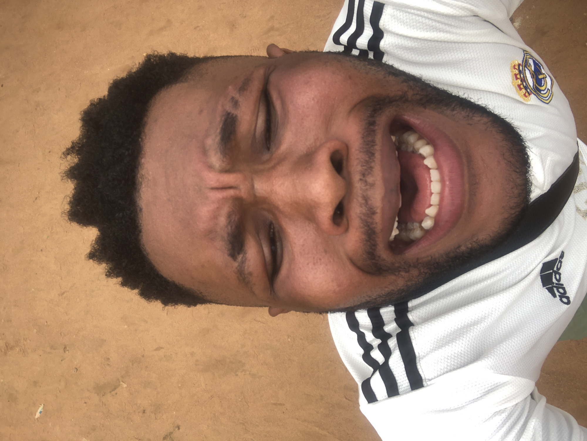 A close-up photograph shows a man in Unknown, Unknown, lying on a dirt surface. The man has dark skin, short black hair, and a beard. His eyes are closed, and he is displaying a wide, toothy grin. He is wearing a white athletic shirt with black stripes on the shoulders and sleeves, featuring the Adidas logo and a Real Madrid emblem. The surrounding environment appears to be sandy or dusty.