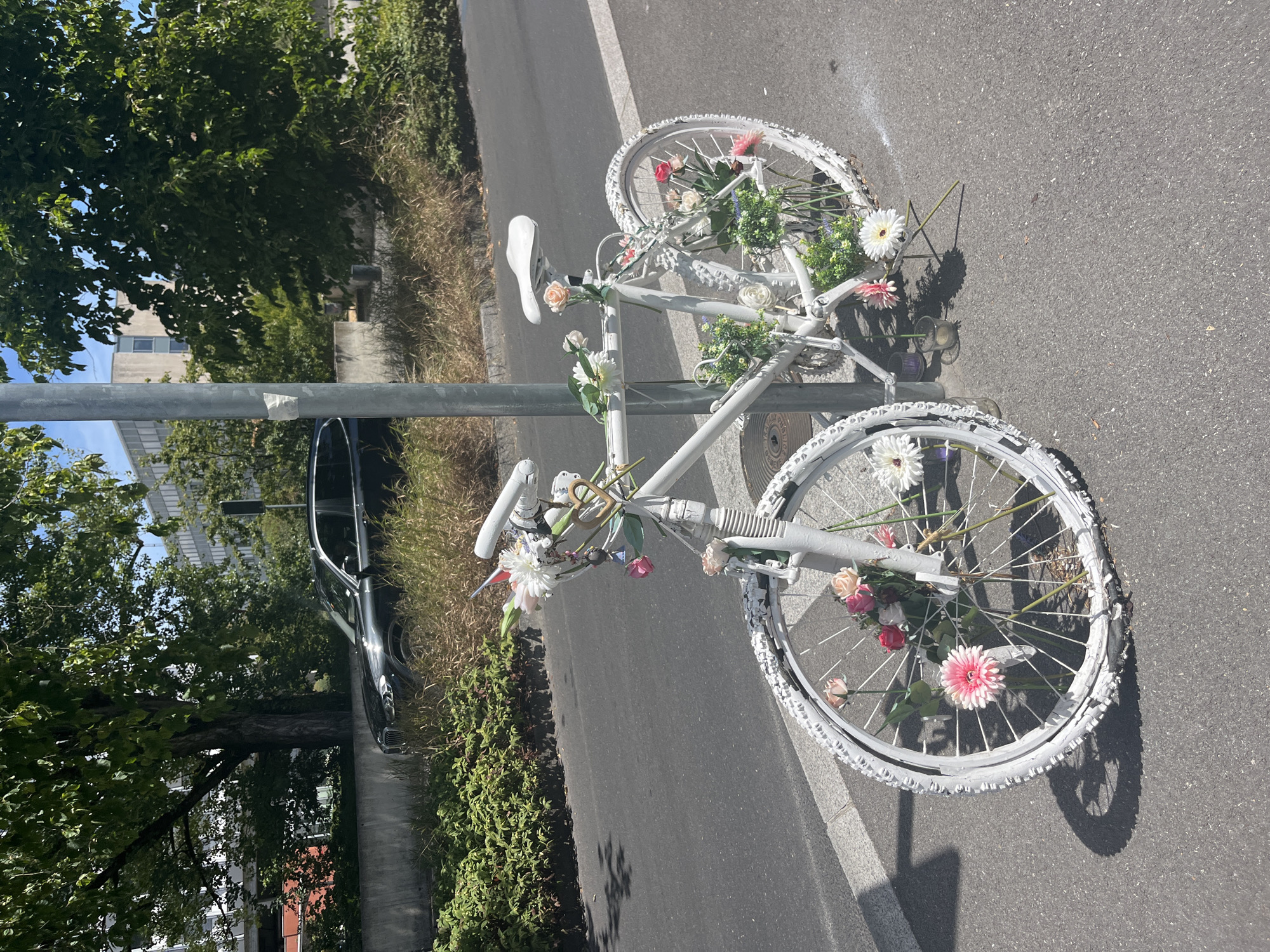 An artistic display featuring a white bicycle decorated with artificial flowers is positioned next to a metal lamppost on a paved sidewalk. The bicycle, painted entirely white, has an assortment of pink, white, and yellow flowers attached to its frame, handlebars, and wheels. A heart-shaped charm hangs from the handlebars, and small flags are also visible. The bicycle is leaning against the lamppost, suggesting it is stationary and part of an exhibit. The scene is set in an urban environment, with a road to the left and a vegetated area beyond that. In the background, a black SUV is parked near a concrete barrier and lush green bushes. Further back, modern buildings with light-colored facades and windows are visible, interspersed with trees. The sky is clear and blue. This scene is located on a sidewalk along the Quai du Mont-Blanc in Geneva, Switzerland.