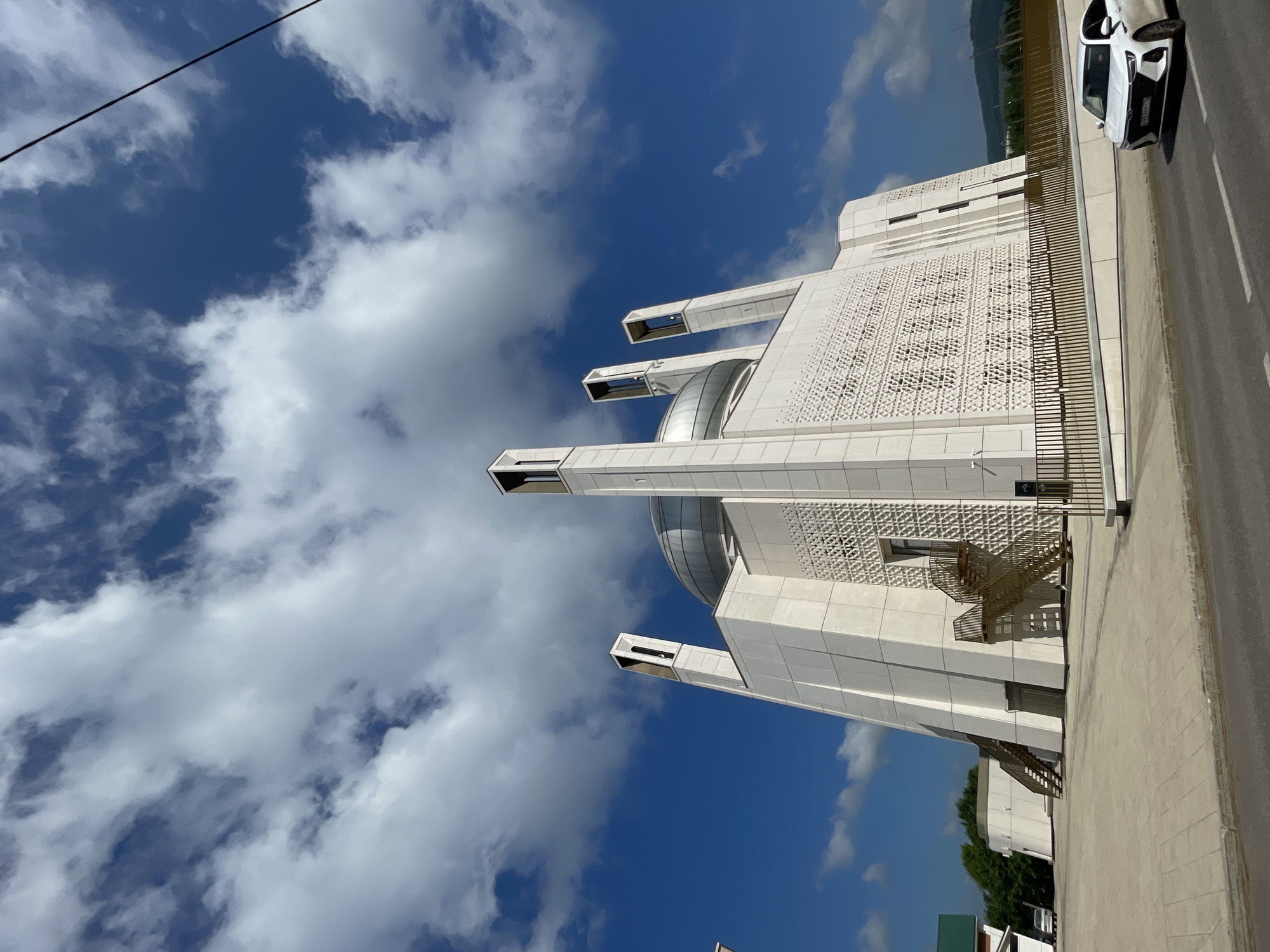 This image captures a modern mosque in Almetyevsk, Russia, under a partly cloudy blue sky. The mosque is a striking white structure with a large central dome and four tall, slender minarets topped with rectangular openings. The exterior walls feature intricate geometric patterns, and external staircases provide access to the building. In the foreground, a white car is visible on an asphalt road. The surrounding environment includes a paved walkway, a fence, trees, and a distant hilly landscape.