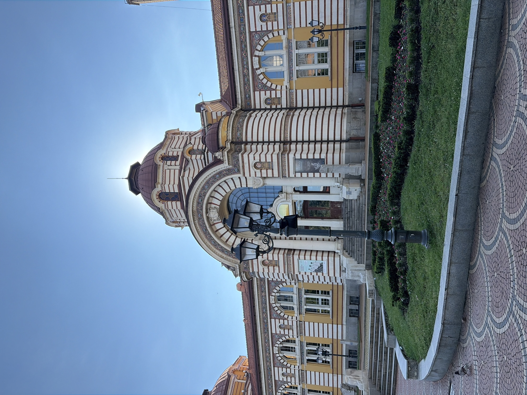 The image shows the Central Mineral Baths in Sofia, Bulgaria, on a clear day with a bright blue sky. The facade of the building is the main subject, characterized by its ornate architecture featuring white and red stripes, arched windows, and a prominent dome. A large, decorative lamppost stands in the foreground, and a neatly manicured garden with flowers and hedges is visible in front of the building. A few steps lead up to the main entrance. A bird is present on the patterned pavement in the lower left corner. The sun is partially visible behind the dome, creating a lens flare effect.