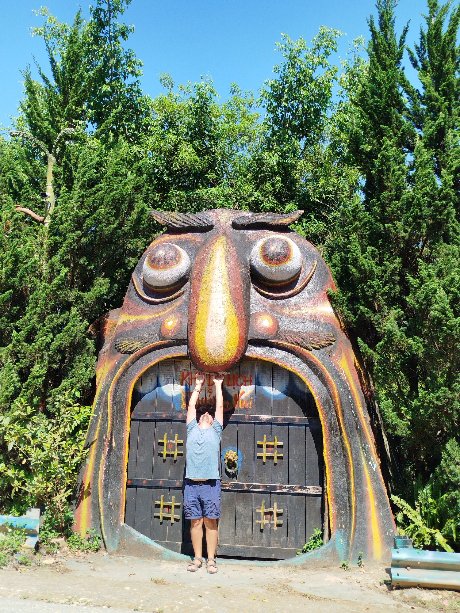 An outdoor, eye-level shot captures a man posing in front of a large, elaborate, monster-like entrance structure under a clear blue sky. The central subject is a towering, stylized head with large, round, yellow and orange eyes, a prominent yellow nose, and a wide mouth forming a dark wooden doorway. The head itself is richly colored in earthy tones of dark brown, black, yellow, and orange, with textured details suggesting feathers or fur.
A single adult male, viewed from behind, stands directly in front of the door. He wears a light blue t-shirt, blue shorts, and sandals, with his arms raised overhead, touching the upper part of the doorway's "mouth," as if holding it open.
The wooden double door features four yellow hash-like symbols and a brass lion's head door knocker on the right panel. Above the man's hands, faint text is visible on the door, appearing to be "KHU DL" followed by partially obscured Vietnamese words such as "LỊCH." This likely translates to "Tourist Area."
The setting is lush and green, with dense foliage and tall, slender conifer-like trees (possibly cypress or juniper) flanking and towering behind the structure. Other broad-leafed trees are also visible. The ground in the foreground is a mix of dirt and gravel, and a blue-green metal guardrail is partially visible on the far right, suggesting a roadside or path. The bright sunlight indicates daytime, casting strong shadows and contributing to a clear and vibrant scene. The man's pose suggests a playful interaction with the whimsical, impressive structure, typical of a themed attraction in a park setting, consistent with the location context of Păng Tiêng 1, Vietnam.