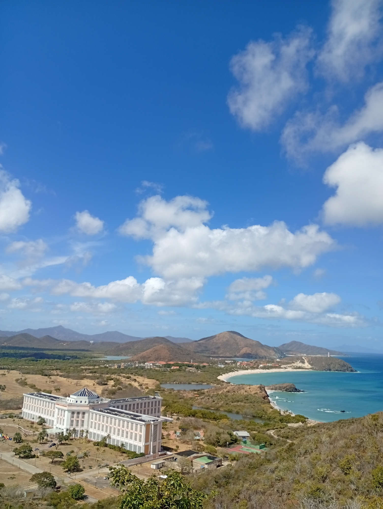 This image displays an elevated, panoramic view of a coastal and inland landscape within Juan Griego, Venezuela, under a clear blue sky dotted with scattered white cumulus clouds. In the foreground, arid, scrub-covered hills slope downwards. The midground features a large, white, multi-story building with a central domed section; the text "HOSPITAL DE LA MARGARITA" is visible on its roof. Paved access roads, landscaped areas, and several parked vehicles surround this structure. To the right, a sandy beach curves alongside turquoise ocean waters exhibiting gentle waves, terminating at a rocky headland crowned by a white lighthouse. Further inland, beyond the main building, the terrain transitions to rolling, arid hills interspersed with natural lagoons and clusters of smaller buildings. No individuals, specific actions, or interactions are discernible.