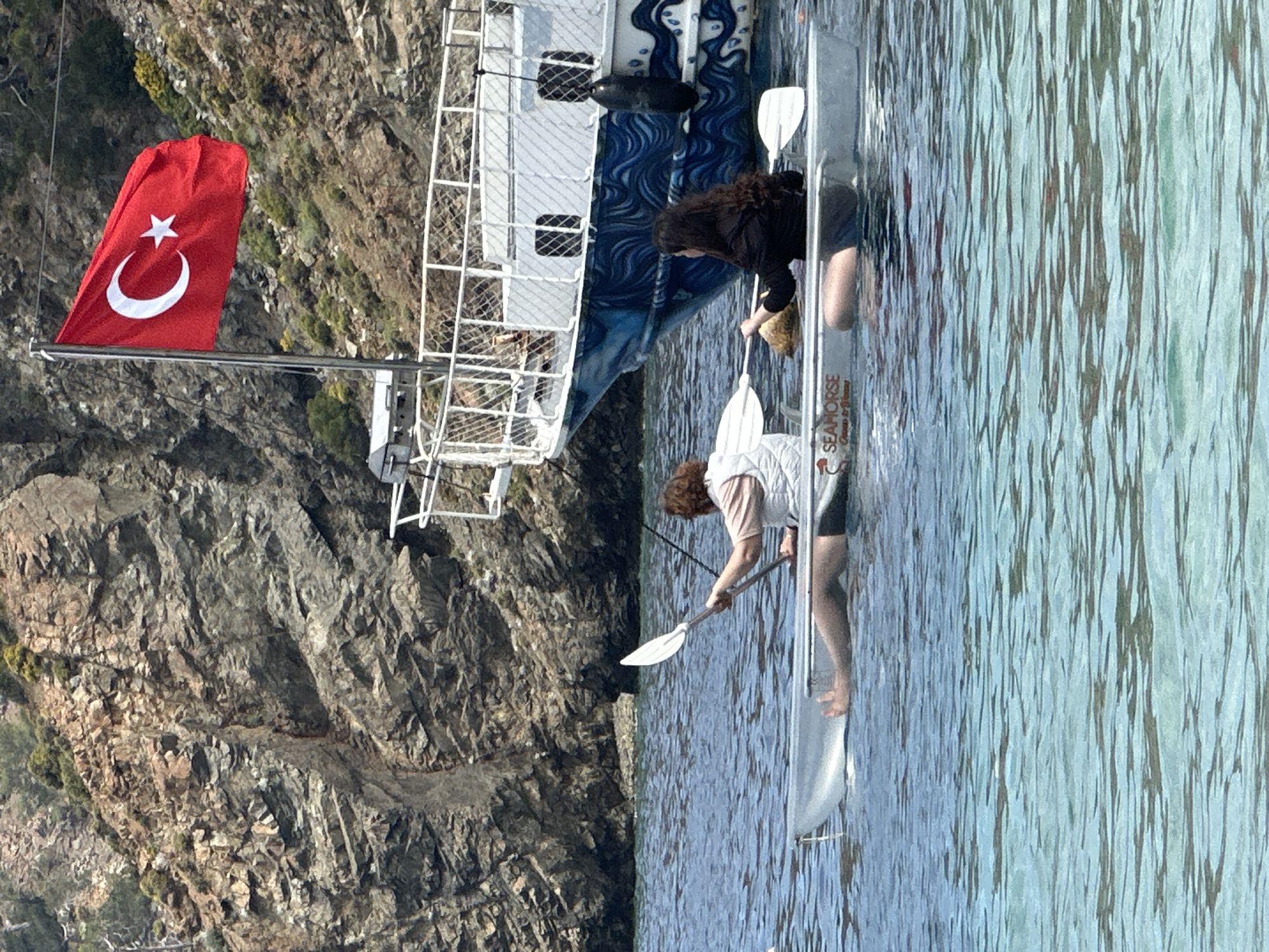 Two women are in a canoe on the water, near a rock wall, on which a Turkish flag is flying. The canoe is next to a ship. One woman is wearing a black shirt and shorts, the other woman is wearing a white shirt and black shorts. The canoe has "Seahorse Cruise & Gulet" on the side. The location is likely within the city of Fethiye, Turkey, due to the presence of the Turkish flag.
