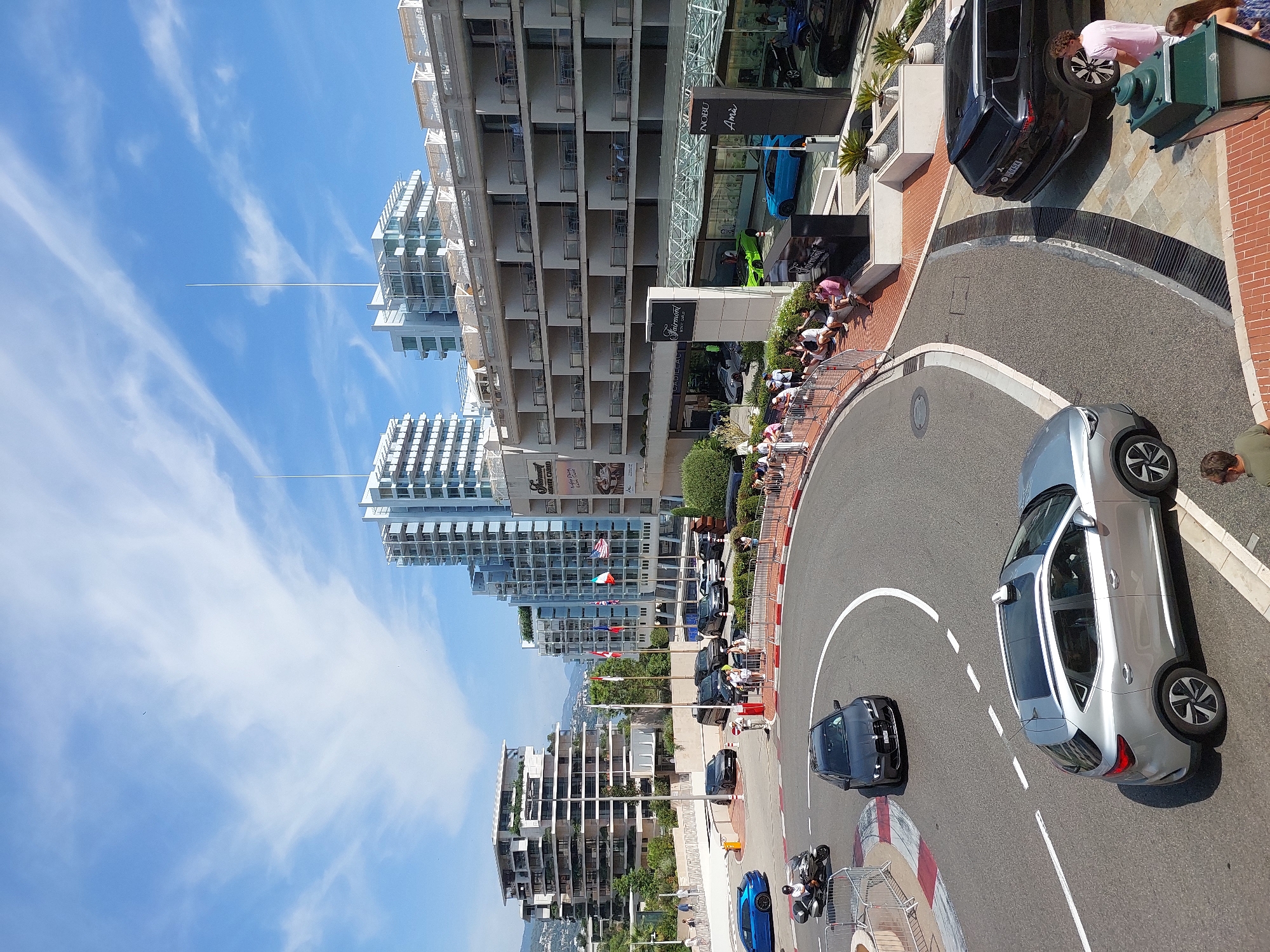The image captures a daytime view of a curved street in Monaco, likely part of the Grand Prix circuit given the barriers and the prominent Fairmont Monte Carlo hotel. Modern, multi-story buildings dominate the background, with a clear blue sky dotted with wispy clouds. In the foreground, a silver SUV with a rooftop antenna is driving on the road, followed by a dark-colored sedan. Further along the curve, a motorcycle with a rider is visible.
To the right of the street, a black SUV is parked near the entrance of the Fairmont Monte Carlo hotel. A group of people are gathered on the sidewalk in front of the hotel, some sitting on a low wall and others standing. The hotel itself features a series of balconies overlooking the street, with some guests visible on them. Several flags are displayed on poles in front of the hotel, including those of Monaco, France, and the United States. Exotic cars, including a bright green one and a blue one, are displayed near the hotel entrance, suggesting a possible car show or event. The overall scene is one of urban sophistication and activity under a bright, sunny sky.