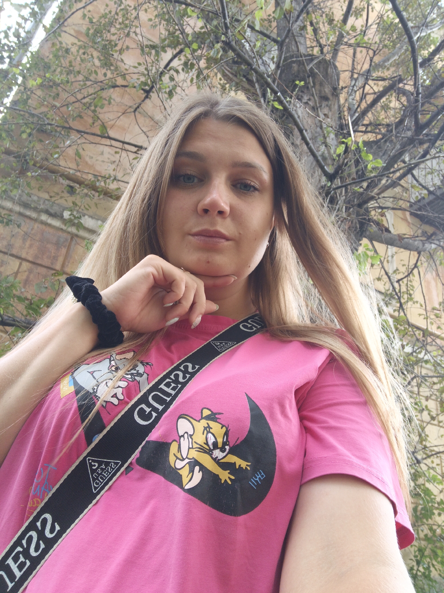A young woman with blonde hair is pictured from the chest up, outdoors, with a blurred background of a building and trees. She is wearing a bright pink t-shirt with graphics of Tom and Jerry characters and a black strap that says "GUESS" across her chest. Her right hand is resting on her chin, and she is looking at the camera. The image was taken on a cloudy day. No specific landmarks of Zlatoust, Russia are visible in this image.