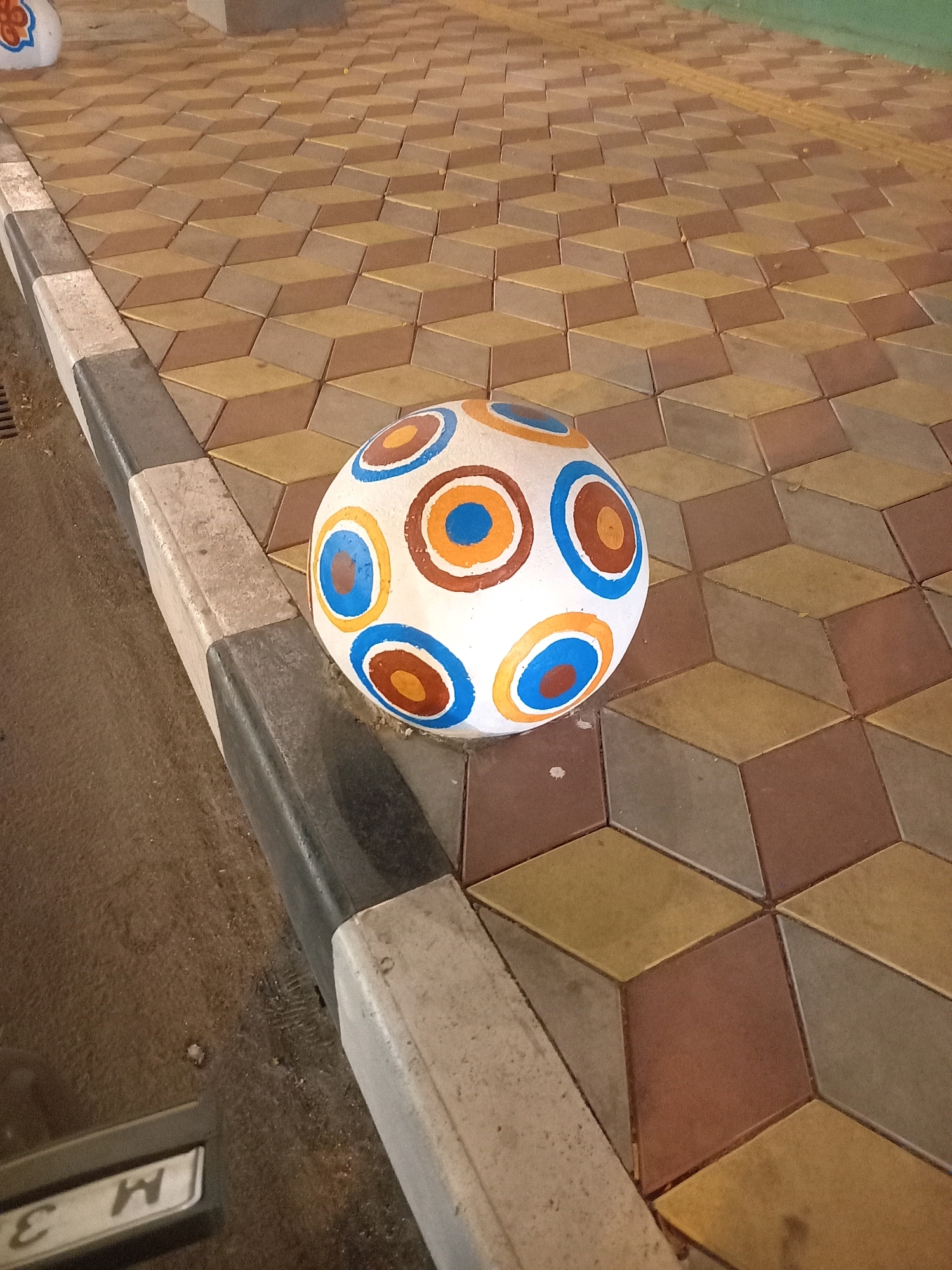 In Pamekasan, Indonesia, a decorative sphere, resembling a soccer ball with concentric circles of blue, orange, and brown on a white background, rests on the edge of a sidewalk. The sidewalk is paved with three-dimensional diamond-shaped tiles in shades of brown, beige, and gray, creating an optical illusion of depth. A curb separates the sidewalk from a dark, textured ground surface, possibly asphalt or dirt. The lower left corner of the image shows part of a metallic object with the letters "M 3" visible, potentially a vehicle license plate or signage. The lighting suggests an outdoor setting, likely during daylight hours.