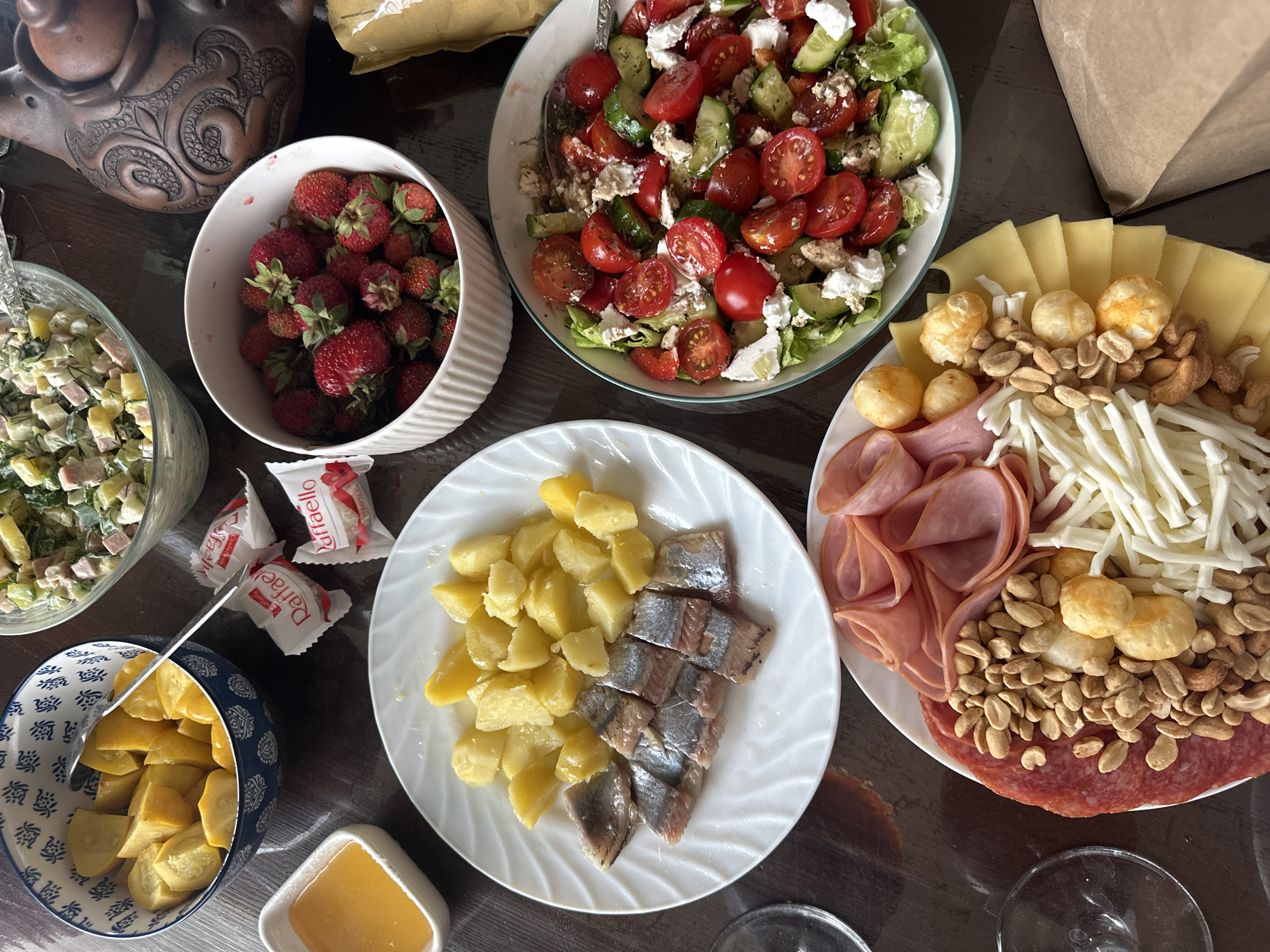 The image depicts a table laden with a variety of food items, suggesting a meal or a gathering. The scene is a close-up, overhead shot of a dark wooden table.
The food includes a bowl of fresh strawberries, a salad with tomatoes, cucumbers, lettuce, and feta cheese, and another salad that appears to be a potato salad with dill and ham. There's a plate with boiled potatoes and pieces of herring, a charcuterie board with sliced salami, ham, various nuts, small cheese balls, and shredded cheese. A blue and white patterned bowl contains chopped yellow fruit, possibly pickled or preserved. A small white dish holds a golden liquid, likely honey or oil. Two wrapped chocolates, identified as "Raffaello" by visible text, are also present. A decorative brown teapot or similar vessel is visible in the upper left corner.
The lighting suggests it is daytime, with natural light illuminating the food. The overall impression is one of abundance and a shared meal. The presence of various dishes and the arrangement of food suggest a home setting, possibly in Chaikovsky, Russia, as per the location context. No people are visible in the image. The focus is entirely on the appetizing spread.