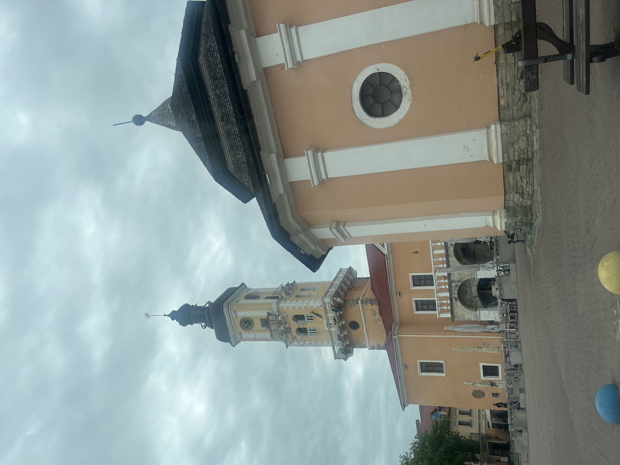 The image captures a view of the Kamianets-Podilskyi Town Hall, a prominent landmark in the city of Kamianets-Podilskyi, Ukraine. The Town Hall features a tall, yellow clock tower with a black spire and a small golden star at its apex. The tower has multiple levels with arched windows and a balcony adorned with a Ukrainian flag. Adjacent to the tower is another building with a peach-colored facade, a pitched roof, and decorative white columns. A circular window is visible on this building's facade. The foreground shows a paved square with several outdoor cafe tables and chairs, umbrellas, and a wooden bench. There are also decorative blue and yellow spheres on the ground. The sky is overcast with clouds.