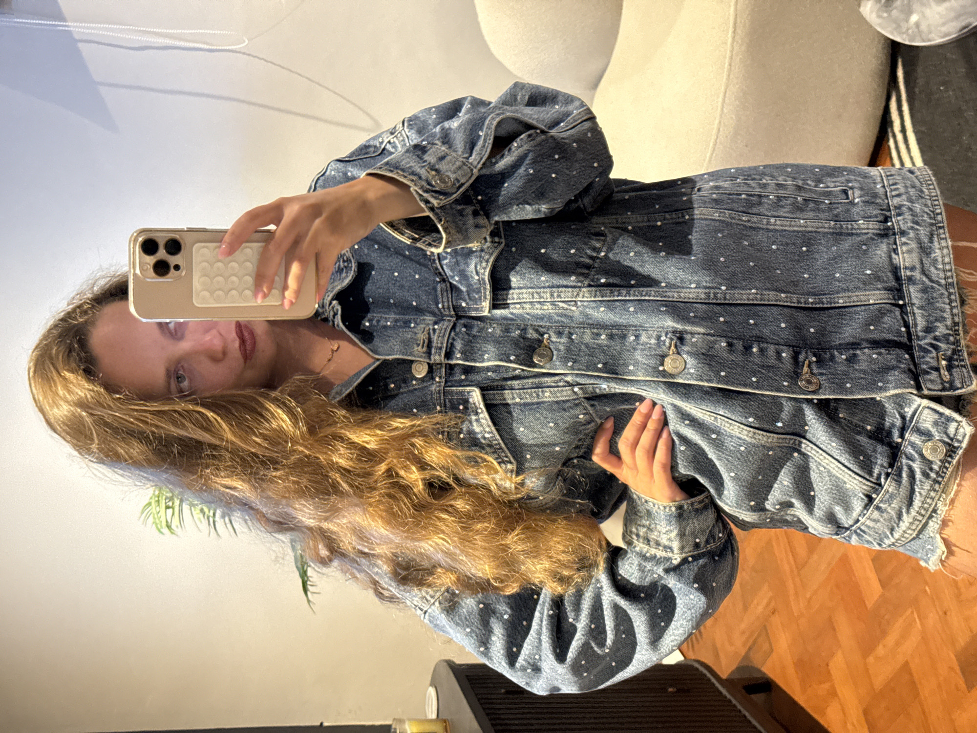 The image depicts a person with long, wavy blonde hair taking a selfie indoors. They are wearing a denim jacket adorned with small, possibly rhinestone, embellishments. The background includes a light-colored sofa, a wooden floor, and a hint of foliage, possibly a houseplant. The scene is likely indoors, but there is no information to place it in Cape Town, South Africa.