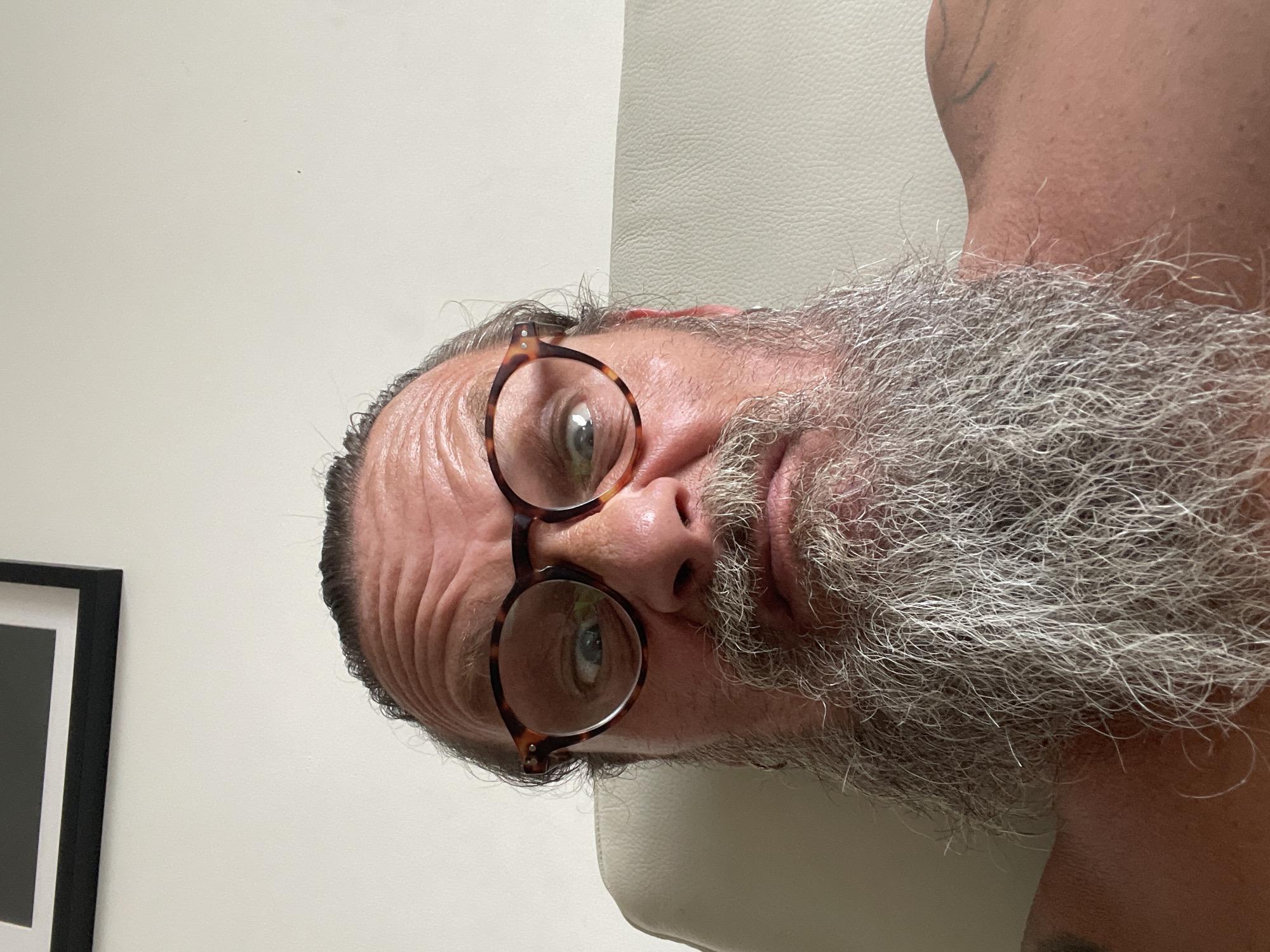 A man with a full grey beard and glasses is looking into the camera. He is sitting on a cream-colored leather chair. The background is a plain white wall with a framed picture hanging on it. The man's eyes are wide and his expression is somewhat surprised. He has wrinkles on his forehead and his hair is pulled back. The image appears to be a selfie. The location is in the city of Netanya, Israel.