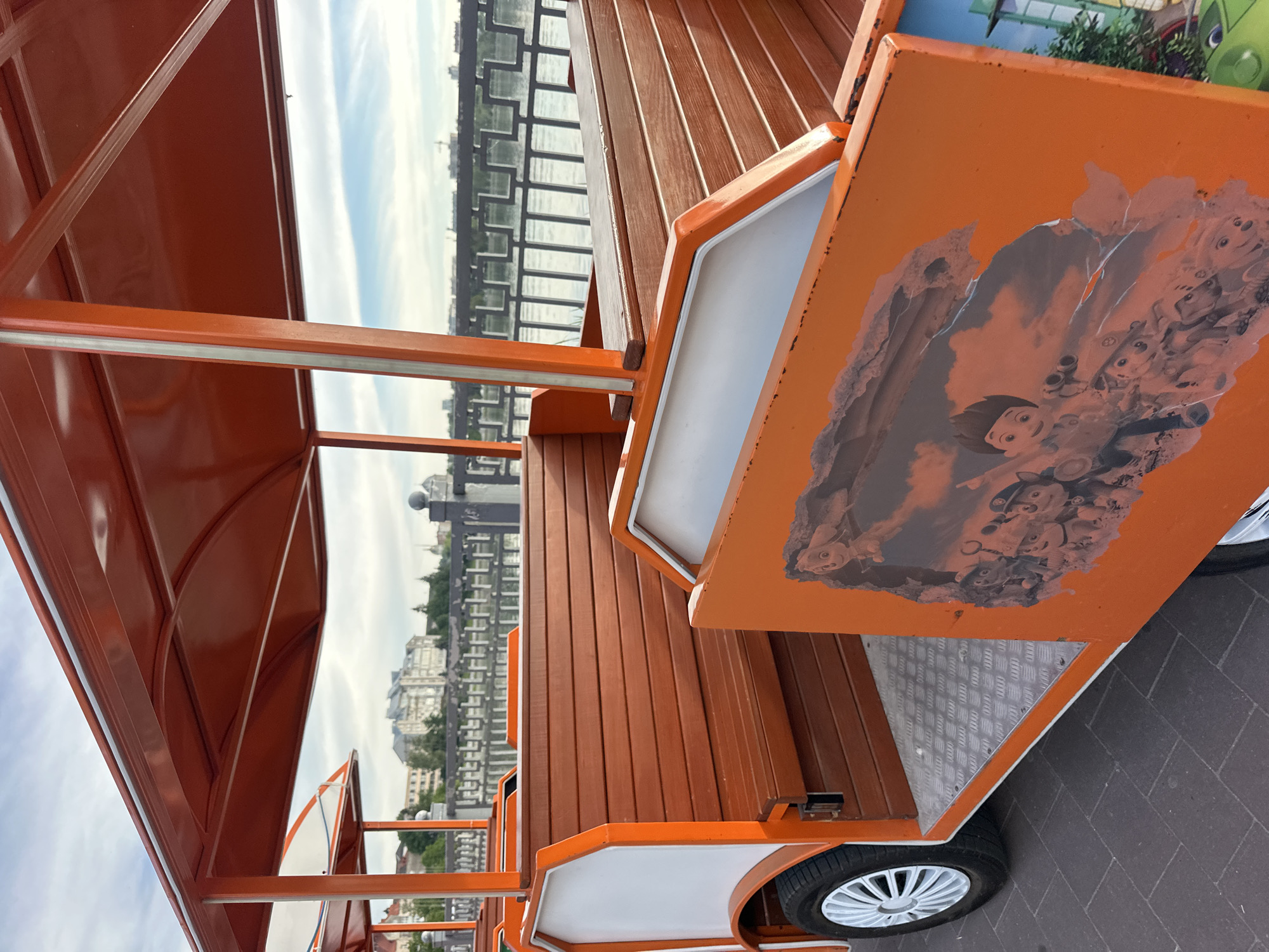 This image shows a close-up of a section of an orange tourist train with wooden benches, parked on a paved walkway next to a body of water with a railing and buildings in the background. The train cars appear to be open-sided, designed for passengers. One car features a partially peeled sticker depicting cartoon characters, possibly from the Paw Patrol series. The sky is cloudy. The location is Ternopil, Ukraine, along the Ternopil Pond promenade.