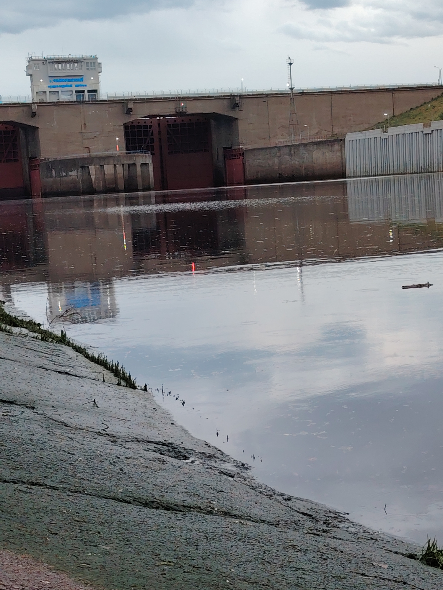The image depicts the Kama Hydroelectric Station in Chaikovsky, Perm Krai, Russia. The shot is taken from a low angle near the water's edge. The hydroelectric station consists of a concrete dam structure with multiple red gates and a control tower building. The tower is white with blue lettering. The water level is high, and the water reflects the sky and the structure of the hydroelectric station. There is a concrete embankment or bank in the foreground. There is a piece of wood floating on the surface of the water. The sky is overcast and cloudy.