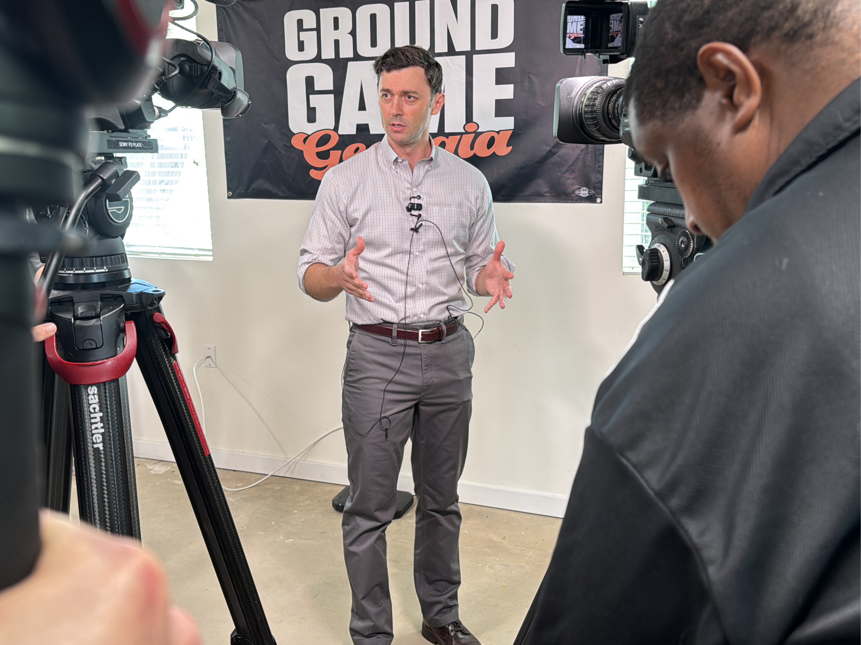 The image depicts a video shoot in what appears to be an indoor studio setting, likely in Decatur, United States based on the "Ground Game Georgia" banner in the background. A man in a light-colored, button-down shirt and gray pants is standing and gesturing with his hands as if speaking or presenting. He is wearing a lapel microphone. Two professional video cameras on tripods frame him on either side; a Black man is partially visible behind one of the cameras. The background consists of a white wall with the aforementioned "Ground Game Georgia" banner. The floor is a light-colored, possibly concrete, surface. Electrical outlets are visible on the wall behind the subject. The cameras appear to be high-end equipment, suggesting a professional video production.