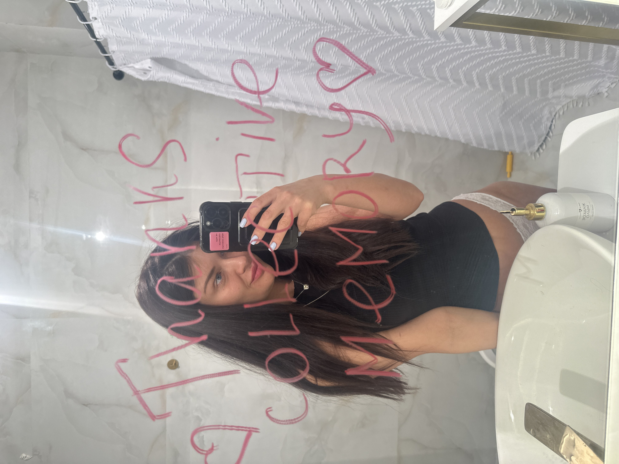 The image displays a mirror selfie taken by a young woman in what appears to be a bathroom, located in Khimki, Russia. The photo is vertically oriented, showing the woman's reflection from the chest up as she holds a black smartphone to capture the image. She has long, dark hair, blue eyes, and is looking towards the camera with a slight smile. She is wearing a black top, with white lace underwear visible above her hip. Her left hand, adorned with light blue nail polish, holds the phone which has a pink sticker on its case.
The mirror's surface is covered with motivational messages and a heart symbol, written in pink lipstick or marker. The text, when read correctly (un-mirrored), says: "SKIN GLOW," "LOVE YOURSELF," "ACTIVE," and "LIVE YOUR BEST LIFE."
In the background, a white shower curtain with a chevron pattern hangs from a rail. To the right of the woman, a white sink and a white soap dispenser with a gold pump are visible. The label on the soap dispenser clearly reads "WIPRO XNAGE." The walls of the bathroom have a light, marble-like pattern. The lighting suggests an indoor setting, and the specific time of day is not ascertainable. The overall impression is a personal moment of self-reflection and empowerment.