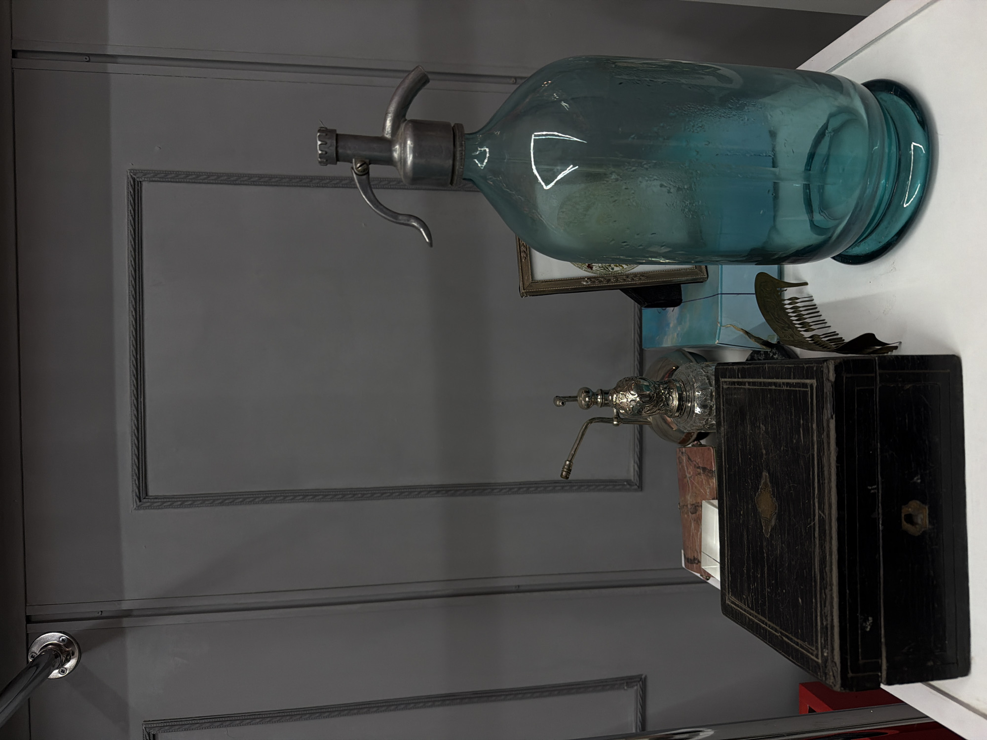 The image displays a still life arrangement of vintage objects on a white surface, positioned against a dark gray paneled wall, within a location in Yerevan, Armenia. In the foreground, a rectangular dark wooden box with a central brass escutcheon and a keyhole is situated on the left. To its immediate right and slightly behind, an ornate silver-colored metallic object, possibly a hookah component, stands. Dominating the right side of the frame is a large, translucent teal-blue glass siphon bottle, showing condensation on its exterior, equipped with a metallic dispensing mechanism on top. A small, gold-framed picture is partially visible behind the siphon bottle. A decorative, antique-style metallic comb lies on the white surface in the mid-ground. The background consists of a uniformly dark gray wall featuring recessed panels with decorative braided trim. A silver-colored metal rail is affixed to the upper left portion of the wall. No people or active interactions are present in the scene.