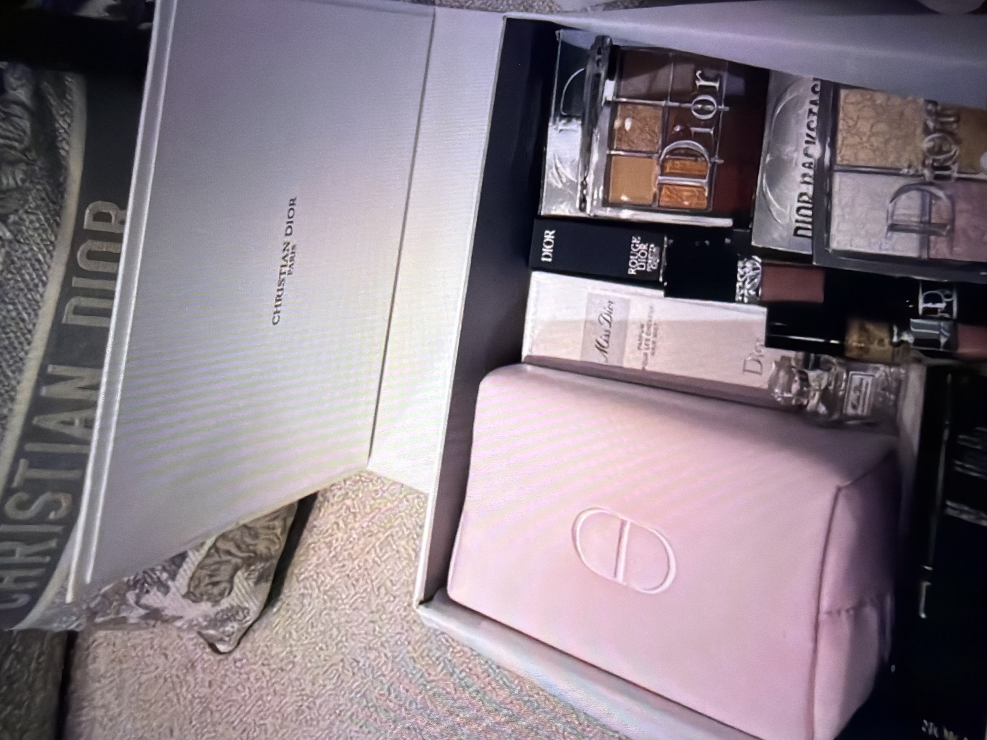 An overhead shot shows a Dior beauty box containing makeup and a makeup bag. The box is open, revealing a pale pink makeup bag with a white "CD" logo on the front. To the right of the makeup bag are several Dior beauty products, including a bottle of "Miss Dior" hair mist, a black tube labeled "ROUGE DIOR," and two eyeshadow palettes. One eyeshadow palette is predominantly brown and gold, while the other contains shades of pink and gold. The box is resting on a textured surface, possibly a carpet or rug. In the background, a pillow with a Dior pattern is visible. The scene is lit, suggesting it is indoors. The image does not contain any visual information that would place it in Beșalma, Moldova.