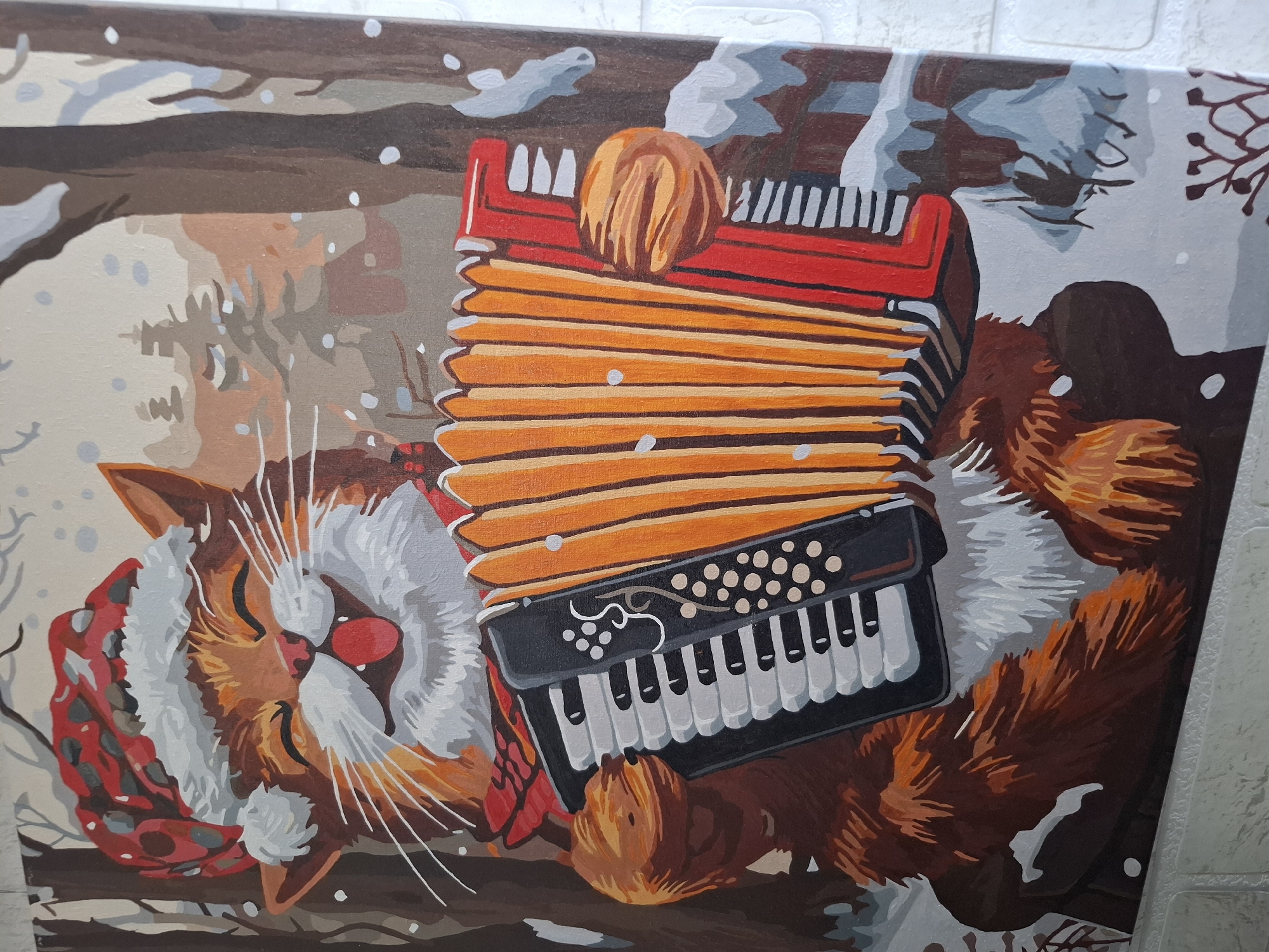 The image shows a close-up of a painting of a cat lying on its back with an accordion on its chest. The cat is orange and white, with its eyes closed. It wears a red and white scarf. The accordion is red, orange, and black. The background is a snowy landscape. This painting is located in the city of Амзя, Russia.