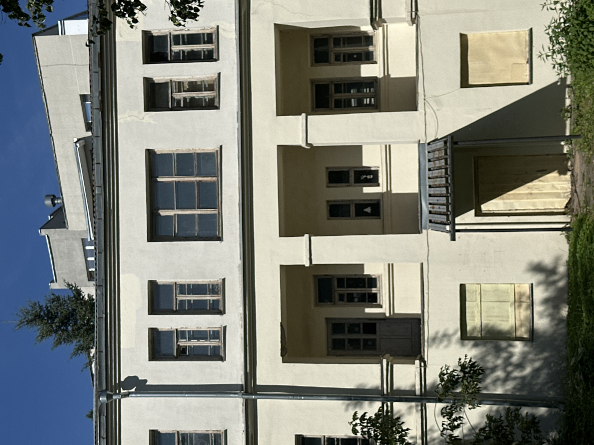 The image shows the facade of a two-story building with a light yellow-colored exterior. The upper story features several large windows with visible wooden frames, some of which appear damaged or weathered. The lower story has a covered porch area with pillars, a doorway, and a window. Below the porch, on the ground level, there are two windows covered with light-colored material. A drainpipe runs down the left side of the building. In the background, other buildings and a tall evergreen tree are visible against a clear blue sky. The building appears to be located in a somewhat overgrown area with grass and foliage in the foreground and shadows cast on the facade. The overall impression is of an older, possibly neglected, urban structure. The building is located in Minsk, Belarus.