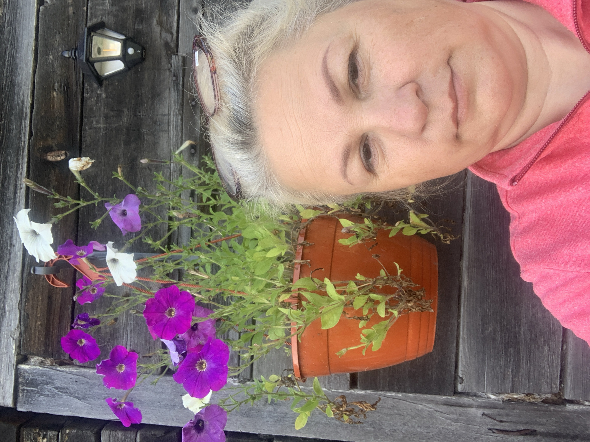 This is a close-up, selfie-style photograph of an adult woman with blonde hair and eyeglasses perched on her head. She is wearing a bright pink zippered jacket and is positioned in front of a weathered wooden wall. To her left, a hanging planter filled with blooming purple and white petunias is attached to the wall. A black outdoor lantern with a light on is visible on the wooden wall behind the flowers. The image appears to be taken outdoors. There is no specific indication of the city of Ust-Ilimsk, Russia, in the visual content of the image itself.