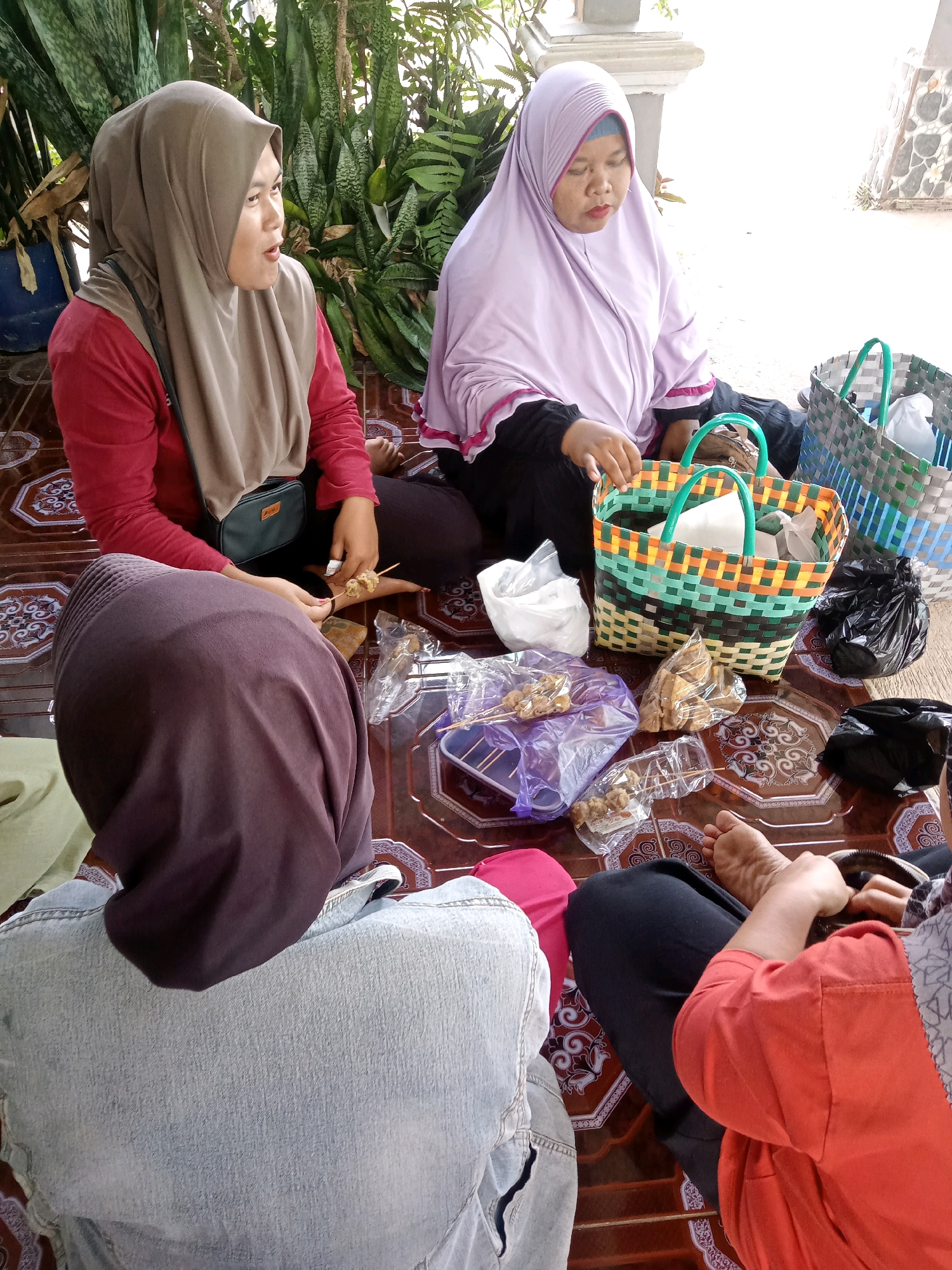 In Gumiwang, Indonesia, four individuals are seated on a patterned tiled floor outdoors, surrounded by lush greenery and a stone structure. The group appears to be engaged in a communal activity involving food. One person, wearing a taupe hijab and a red shirt, holds a skewer with food items. Another person, in a light purple hijab, is seated beside a colorful woven basket filled with items. A third person, facing away from the camera, wears a dark brown hijab and a denim jacket, while the fourth person, wearing an orange top, is partially visible on the right side of the frame. Several food items, including skewers and packaged snacks, are scattered on the floor, along with plastic bags and woven baskets. The scene suggests a casual gathering or meal shared outdoors.