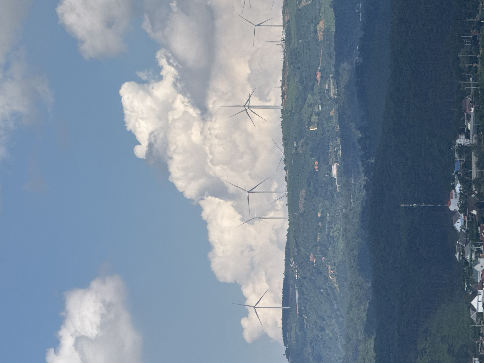 The image shows a landscape with wind turbines on a hill overlooking a town. The sky is blue with white clouds. The hill is covered with green vegetation and some buildings are visible. In the foreground, there is a cluster of houses. The wind turbines are tall and white, with three blades each. They appear to be in operation, as their blades are in motion. The town in the foreground consists of several houses with red and white roofs, and a tall communication tower. The scene suggests a rural or semi-rural setting in Đa Lộc, Vietnam, where wind energy production is likely taking place. The overall atmosphere is peaceful and serene.