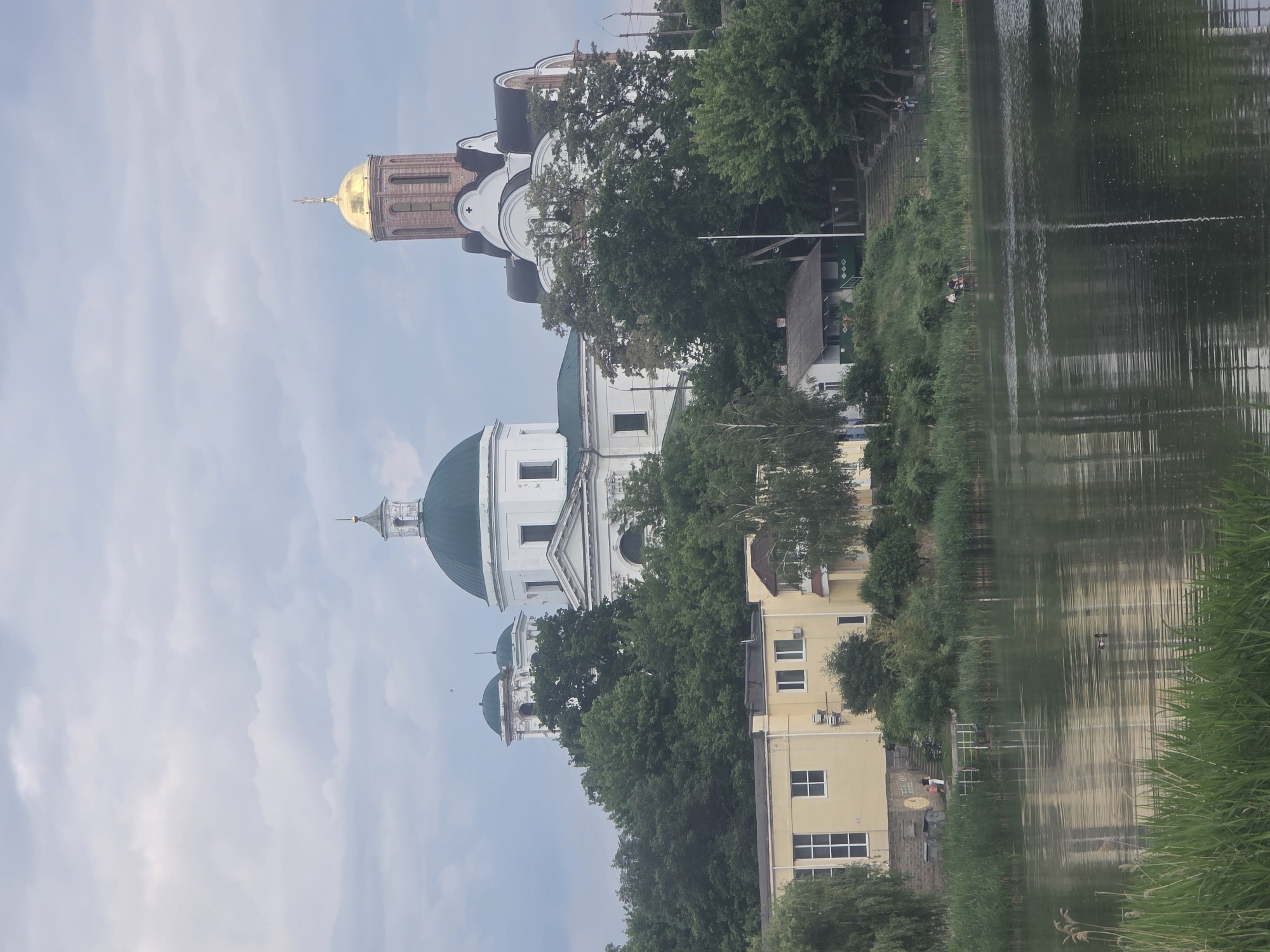 This image presents a scene in Bila Tserkva, Ukraine, featuring prominent religious architecture and natural elements viewed across a body of water.
In the background, under an overcast sky, two cathedrals are visible atop a rise. On the left, a large white structure with a dark green dome and a smaller white cupola topped with a spire stands prominently, identifiable as St. Nicholas Church (Mykolaiv Cathedral). To its right, a tall, multi-tiered brick tower culminates in a golden dome adorned with a cross, indicative of the Transfiguration Cathedral. This tower is integrated with a lower white building featuring dark, multi-faceted roofs. Dense green deciduous trees partially obscure the lower sections of these cathedrals and cover the surrounding hillside.
In the midground, situated on the bank below the cathedrals, a multi-story yellow building with multiple windows and exterior air conditioning units is visible on the left. Further to the right and lower on the bank, a smaller white house with a dark roof is partially visible amidst the trees.
The foreground is dominated by a calm body of water, likely the Ros River, which exhibits subtle ripples and reflects the sky and surrounding vegetation. The immediate bank is lined with tall green reeds and grasses. On the left side of the bank, near the yellow building, two individuals—one adult and one child—stand on an embankment adjacent to a small, brightly colored pier or railing extending into the water. Further to the right along the water's edge, two additional individuals are seated on a low structure. A single dark-feathered duck swims in the water in the middle-right section of the frame. Concrete steps are visible ascending the vegetated bank on the right side of the image, with several individuals dispersed along them or the upper bank.
