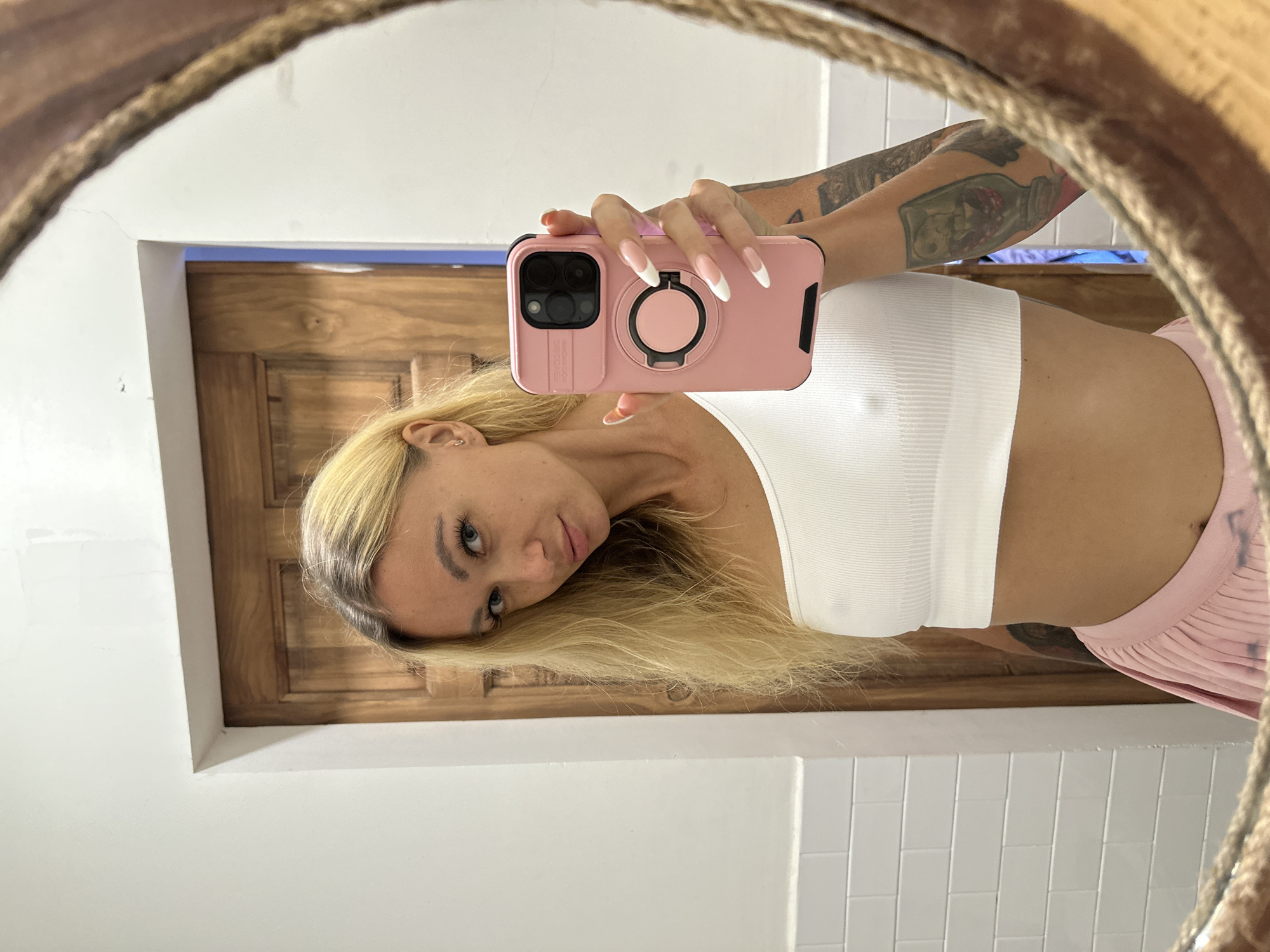 This is a mirror selfie taken by a young woman with blonde hair. She is wearing a white ribbed tank top and a pink pleated skirt. Her right arm is extended, holding a pink phone case with a ring attached. The phone case has a black camera cutout. She has long, sculpted fingernails painted with a French manicure. Tattoos are visible on her upper right arm. The background shows a wooden door and white tiled walls. The mirror has a thick, braided rope frame. The lighting suggests it is daytime, and the setting appears to be indoors, possibly a bathroom or bedroom, given the tiled walls and the mirror. The woman's expression is a slight smirk, and her head is tilted, giving the photo a playful feel. There is no visible text in the image.