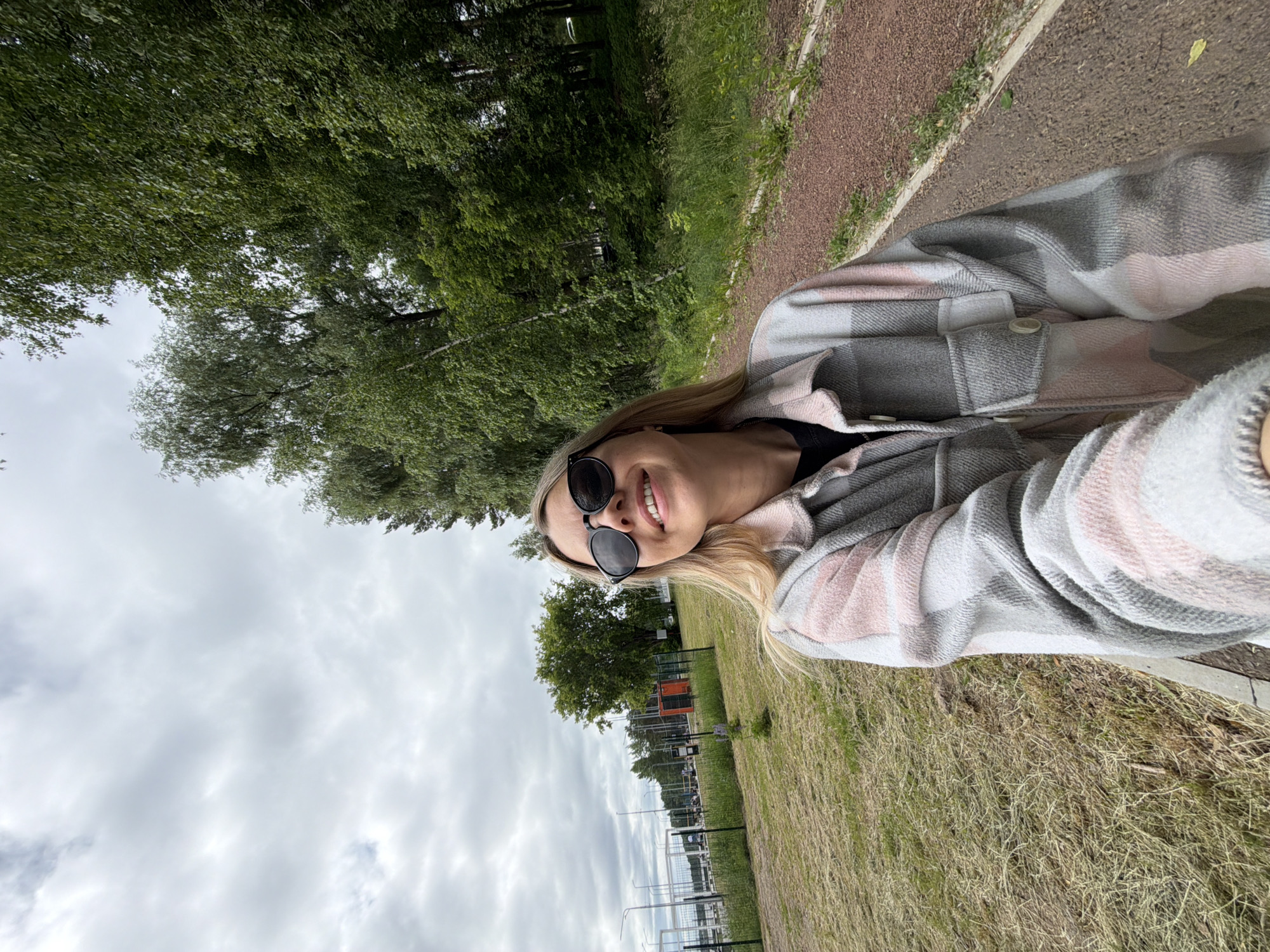 In Chaikovsky, Russia, a young woman with blonde hair and sunglasses is taking a selfie outdoors. She is wearing a plaid shirt with pink and gray colors over a black shirt. She is smiling and looking up. The background shows a walking path made of reddish gravel, a grassy field, and trees. In the distance, there are sports facilities and streetlights. The sky is cloudy.