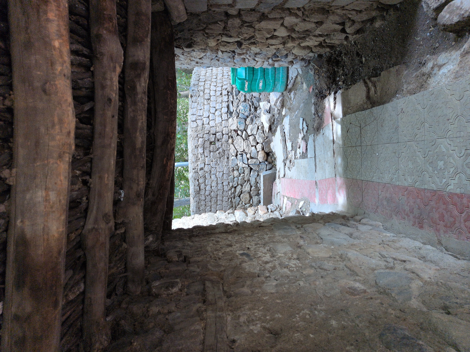 The image shows a narrow passage with rough stone walls on either side, leading to an outdoor area. The passage has a tiled floor with a red and gray pattern. Overhead, rough-hewn wooden beams form a rustic ceiling. The outdoor area beyond the passage features more stone walls, a large green water tank, and scattered debris. The scene appears to be in a historic or traditional setting, possibly within the city of Leh, India, given the architectural style and materials used. No people are visible in the image.