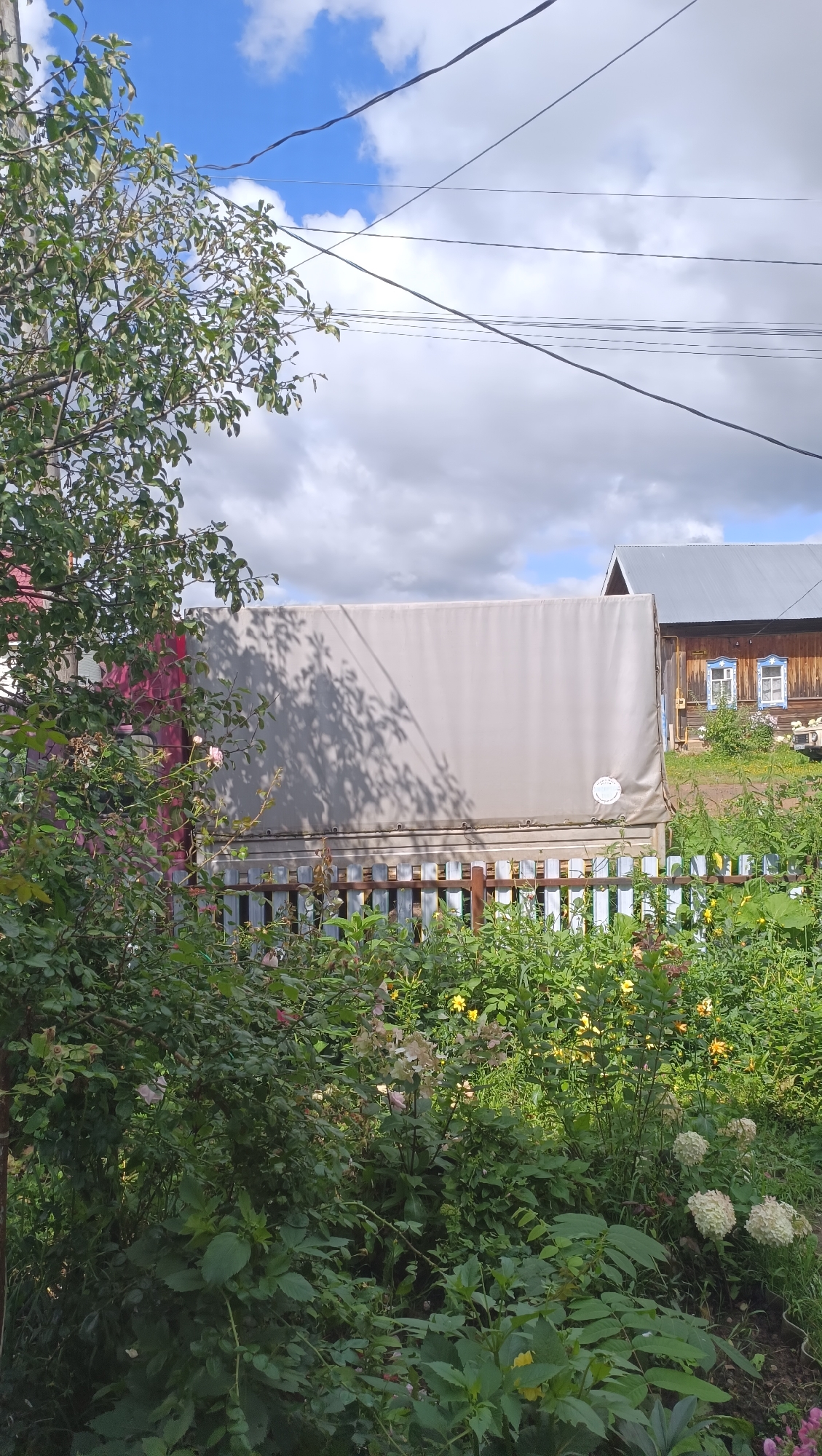 In the city of Karsha, Russia, a daytime scene unfolds with a wooden house and a covered vehicle. The house, with a gray roof and wooden siding, features two windows with blue trim. In front of the house, a light-colored canvas-covered truck is parked, its side partially obscured by a white picket fence and lush green vegetation. The foreground is filled with dense foliage, including flowering plants and bushes, some with pale pink blossoms. Overhead, a bright blue sky is dotted with fluffy white clouds, and power lines crisscross the upper portion of the frame. The sunlight casts dappled shadows from tree branches onto the canvas of the truck.