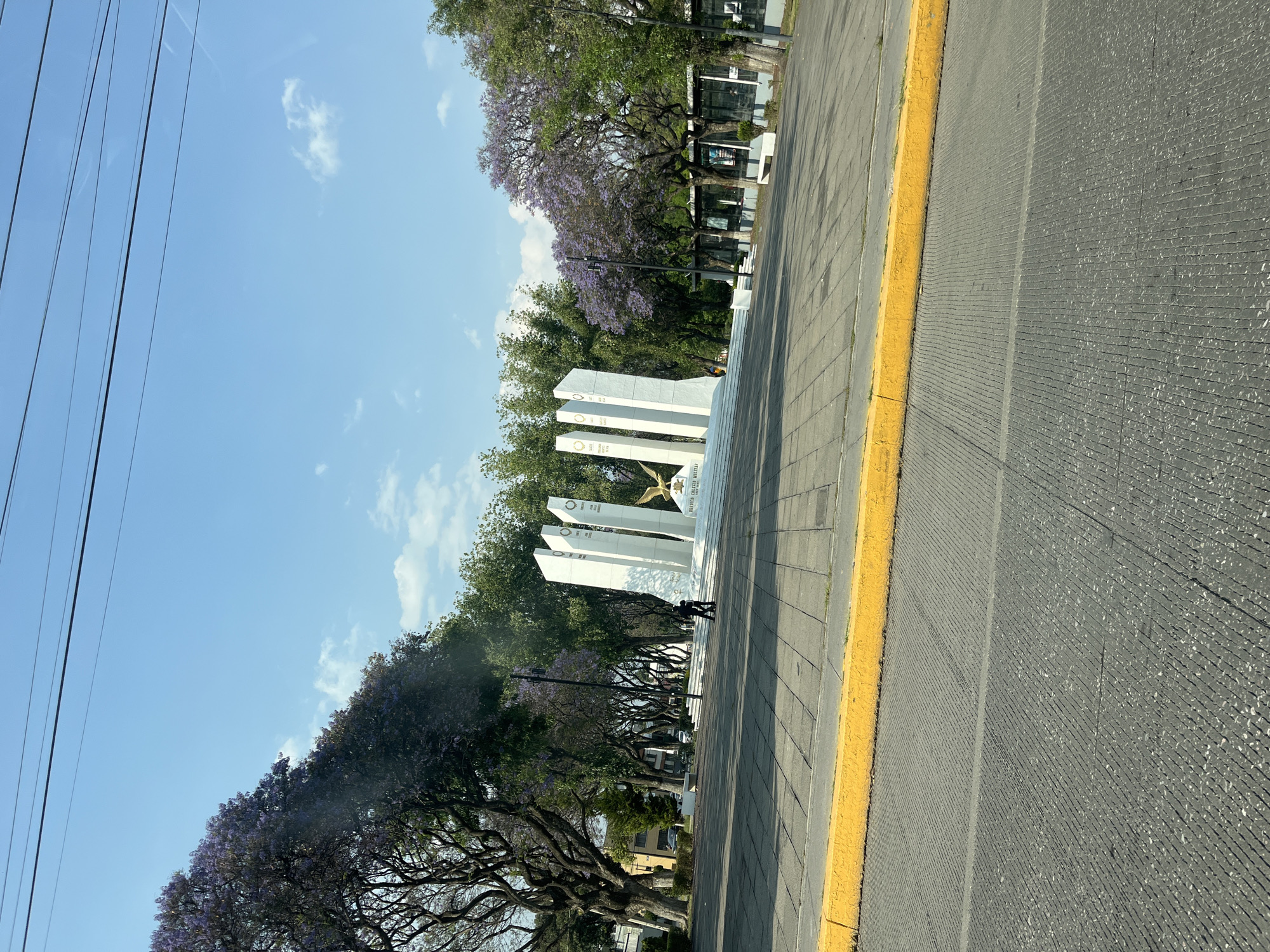 A daytime, eye-level shot shows a street scene in Puebla, Mexico. The camera is angled, creating a tilted perspective. On the left, electrical lines run along the sky above trees with purple blossoms. A monument consisting of white pillars and a gold statue is on the sidewalk. The sidewalk, paved with gray bricks, has a yellow curb separating it from the street. Two indistinct figures are walking in front of the monument. The background includes green trees and glimpses of buildings. The sky is blue with a few white clouds.