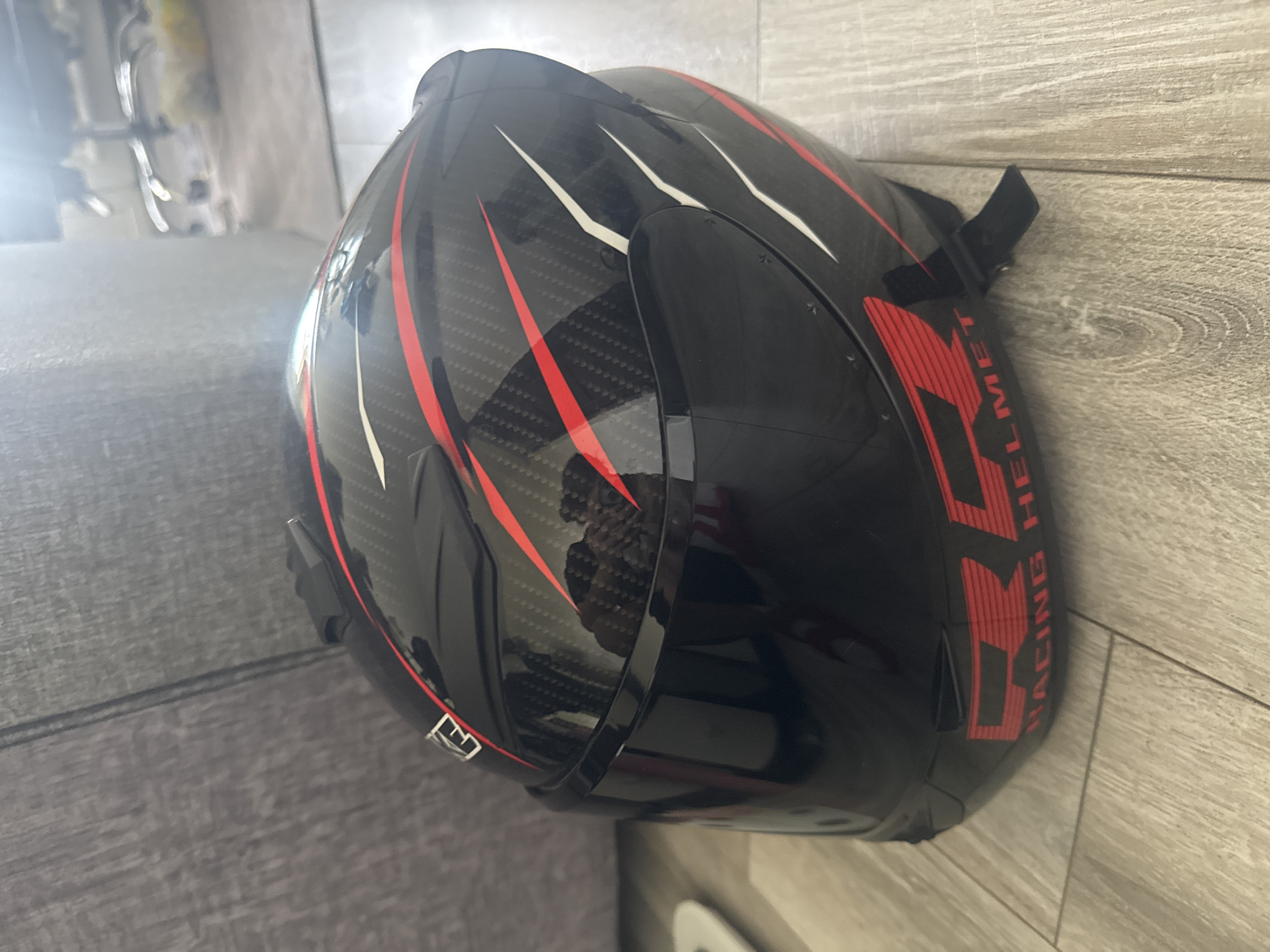 A black motorcycle helmet with red and white accents and a carbon fiber pattern is shown in a close-up shot. The helmet has a clear visor and a chin strap. The helmet is resting on a light-colored wooden floor. In the background, a gray textured surface and blurry office chairs are visible. The image appears to be taken indoors in an apartment or home setting.