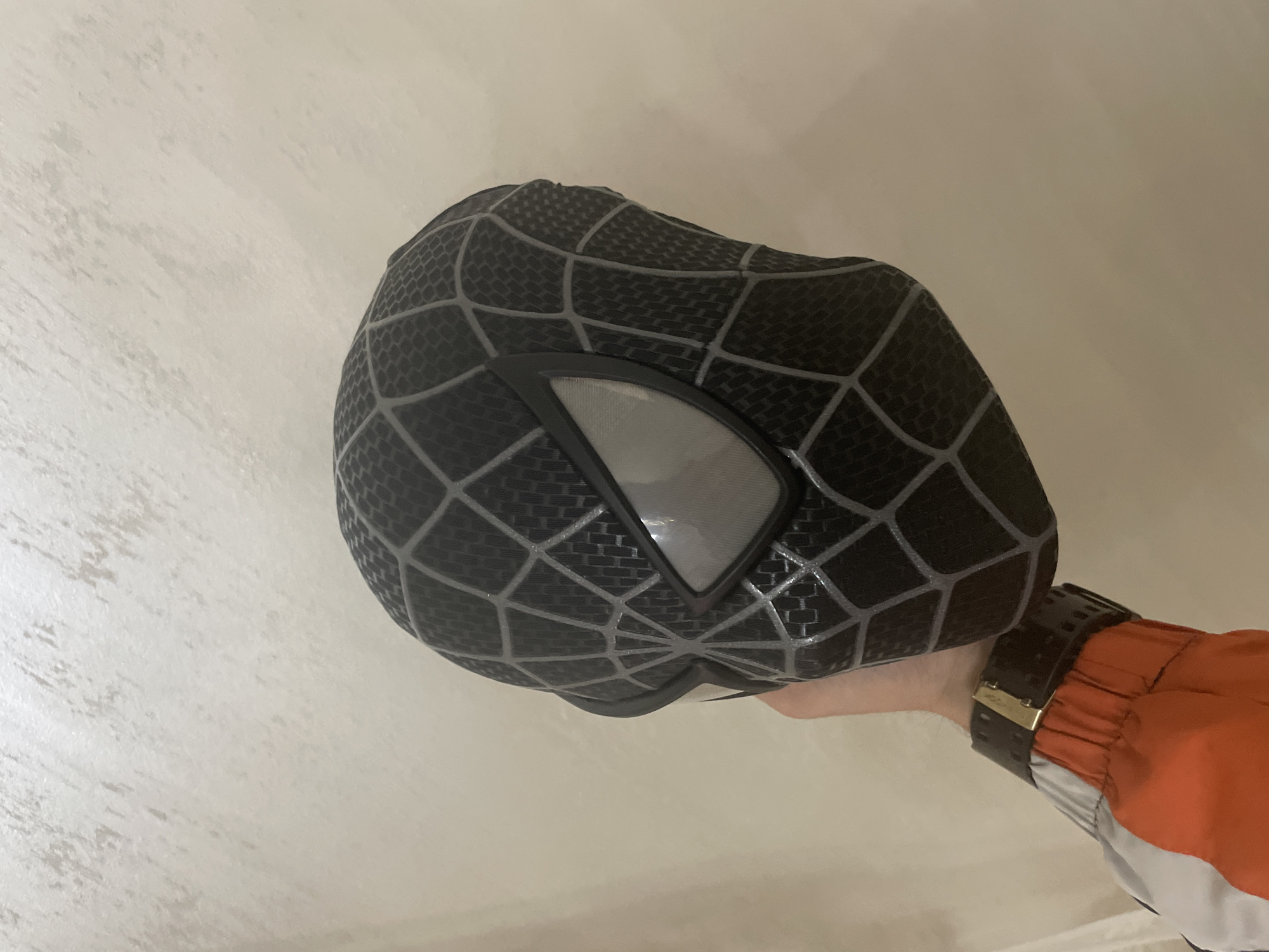 A black Spider-Man mask, featuring a prominent gray web pattern over a black textured surface, is held by a left hand in an indoor setting. The mask's eye lenses are large, reflective, and silver-toned, framed by black borders. The hand, with fair skin, supports the mask from below. A dark-colored, wide wristband with a gold-toned buckle and perforated detailing is present on the wrist. The sleeve of a multi-colored garment, predominantly orange with a white section, is visible on the arm, displaying an elasticated cuff. The background consists of a light-colored wall with an irregular, textured finish. The scene is located in the city of Ihtiman, Bulgaria.
