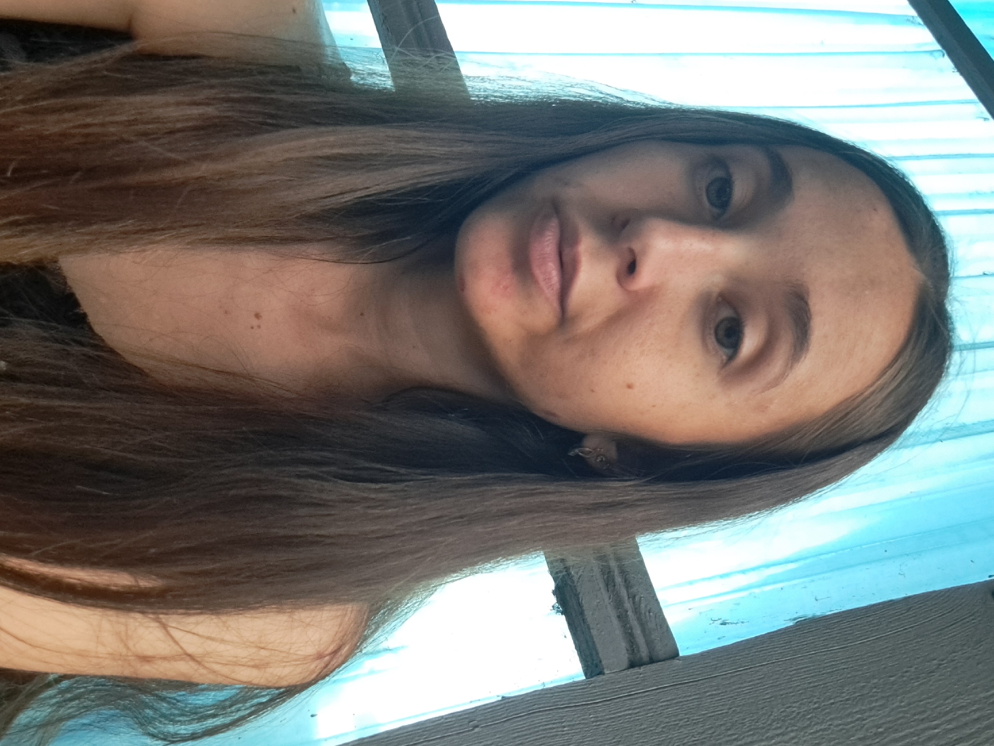 A close-up, medium shot shows a young woman with long, dark brown hair framing her face, which is looking directly at the camera. She has fair skin with some freckles and minor blemishes on her chin. She is wearing a dark-colored top. The background consists of vertical blue plastic sheeting and a gray wooden structure on the right side of the frame. The lighting suggests an outdoor setting, possibly a porch or covered area, with sunlight filtering through the blue material. The scene is static and appears to be a selfie taken in Vypasne, Ukraine.