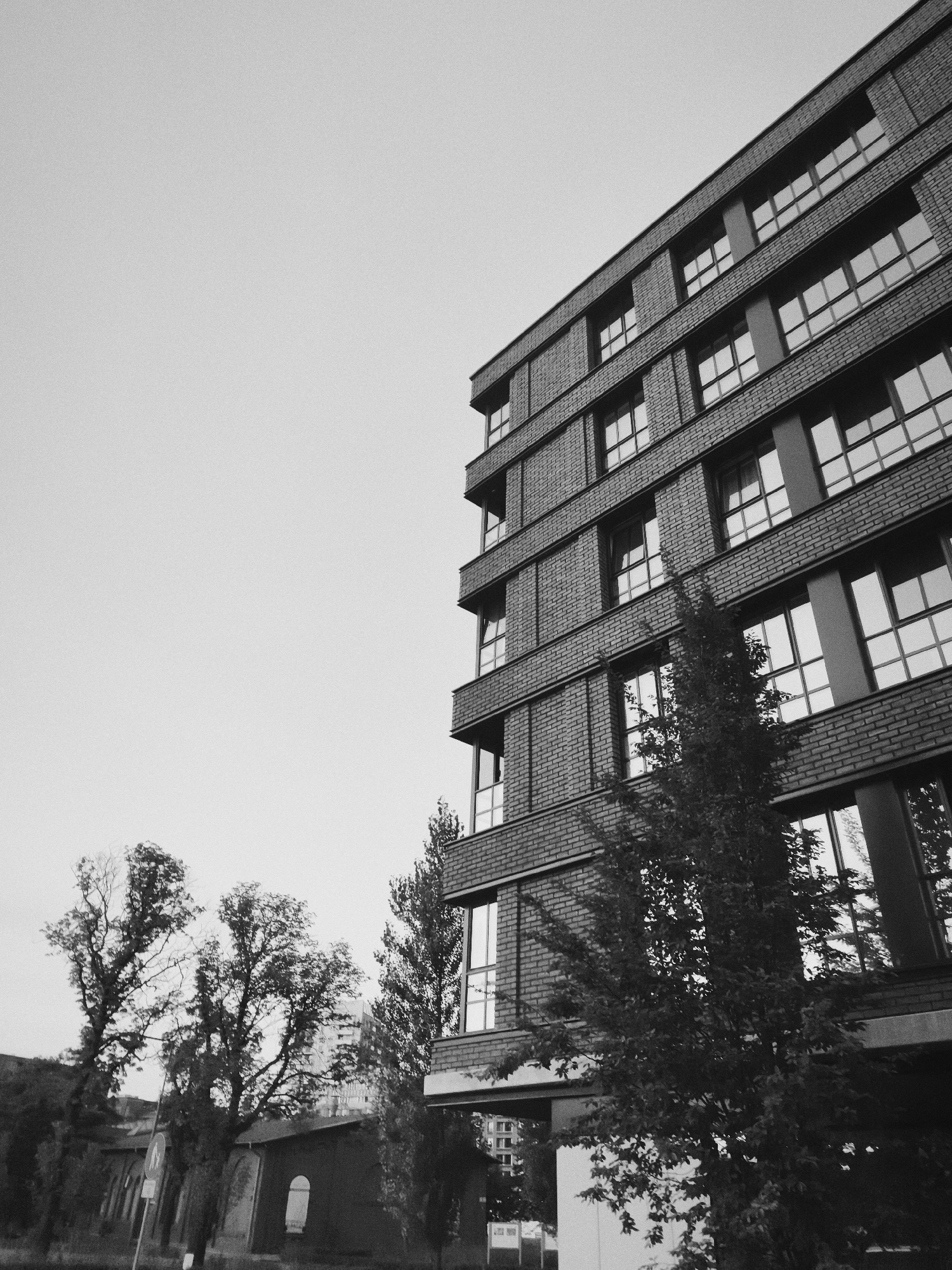 The image, captured in black and white, depicts the exterior of a modern brick building with numerous windows. The building is angled, with the top right corner of the frame showing a section of its facade. Trees, some bare and others with foliage, are visible in the foreground and to the left of the building, casting shadows. The sky is overcast and light. The scene appears to be in an urban environment, possibly in the city of Gdańsk, Poland, judging by the architectural style and the presence of other buildings in the background. No people are visible in the frame.
