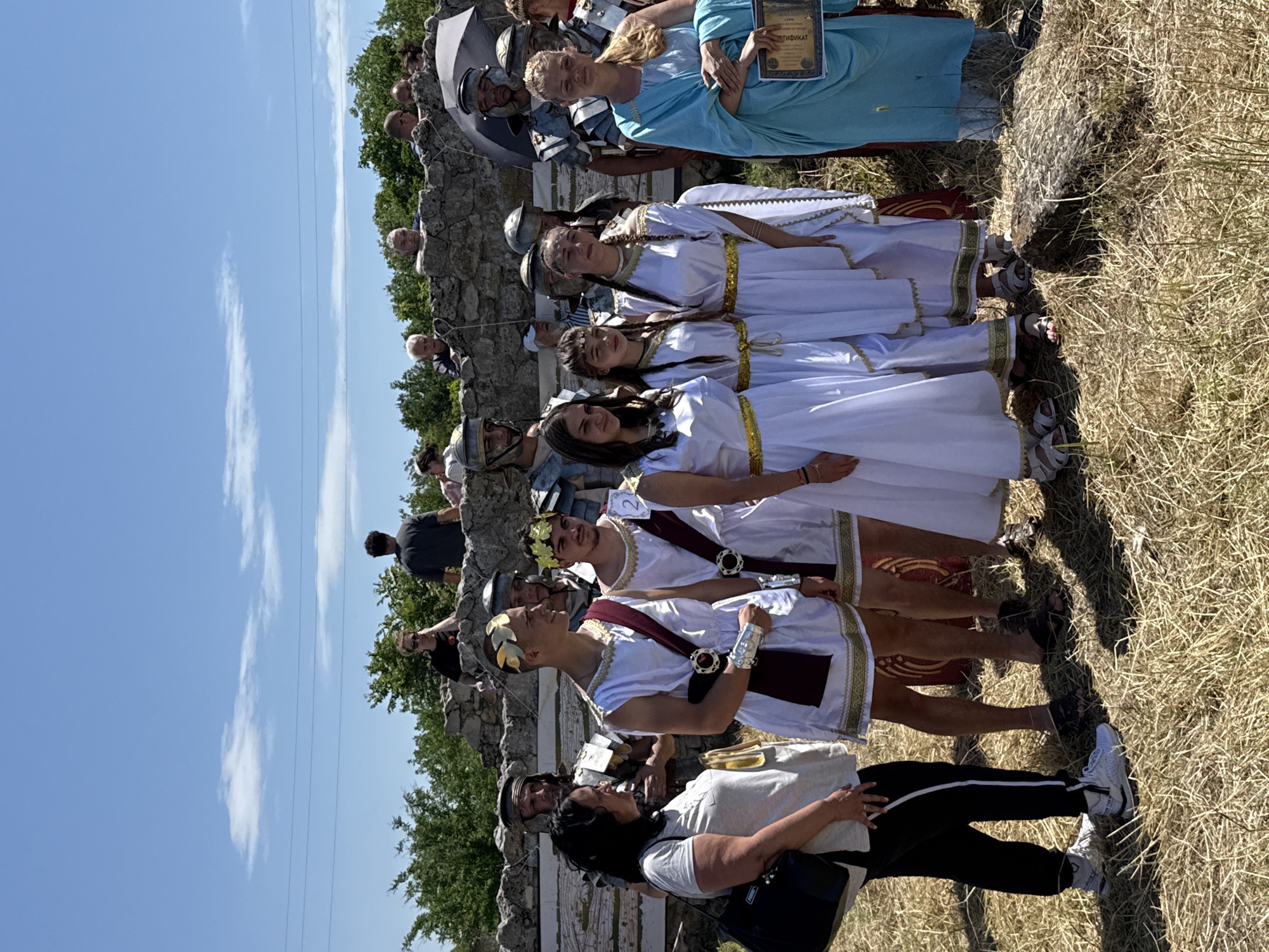 The image depicts an outdoor historical or cultural event at the archaeological site of Oescus in Gigen, Bulgaria, under a clear blue sky with sparse clouds. In the foreground and mid-ground, approximately eight individuals are dressed in historical costumes. Two young males wear white tunics with maroon sashes and gold trim, one featuring a gold laurel wreath and silver armbands, the other a gold laurel wreath. Three young females wear white dresses with gold trim. An adult female wears a light blue flowing garment and holds a framed certificate or diploma with Cyrillic text. Behind these costumed figures, several men in Roman legionary armor (helmets, breastplates, shoulder armor) are visible. To the far left, an adult female in modern attire (white t-shirt, black track pants, white sneakers, black handbag) stands, observing the costumed group. In the background, atop and behind a low, irregularly constructed stone wall, numerous individuals in contemporary casual clothing are observing the scene. The ground is covered with dry, yellowish-brown grass and sparse vegetation. Green trees and bushes are visible beyond the stone wall, and faint horizontal power lines traverse the upper background sky. The scene is brightly lit by sunlight, casting visible shadows.
