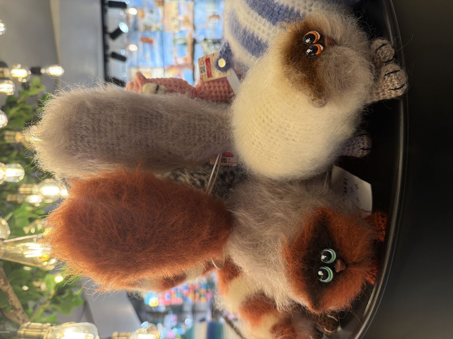 This vertically oriented close-up image showcases a collection of fluffy, knitted or crocheted animal-like figures, likely toys or keychains, displayed in what appears to be an indoor retail environment in Saint Petersburg, Russia.
The foreground features multiple figures. One prominent figure is light grey with a shaggy, fuzzy texture. Below it, a reddish-orange figure with an equally soft, textured surface is visible. To the right, another distinct figure is cream or off-white with brown markings around its face, possessing large, reflective eyes with black pupils and orange irises. This figure also has small, knitted paws. Further down, a fourth figure, reddish-orange and white, is seen with similar large, expressive eyes featuring black pupils and green irises. The overall texture of these items is notably soft and fuzzy, indicating their material is a delicate, fine yarn.
The figures are arranged closely together, suggesting they are part of a product display. In the blurred background, numerous warm-toned light fixtures, resembling Edison bulbs, are visible, creating a soft, luminous effect. Hints of other merchandise or display structures can also be discerned, reinforcing the setting as a shop or market stall. Small white tags with unreadable black text, possibly Cyrillic characters given the location context, are attached to some of the figures. A red tag with the number "100" is also partially visible on one item. No human activity is directly depicted, but the arrangement implies items available for purchase or browsing. The scene conveys a charming and whimsical aesthetic.